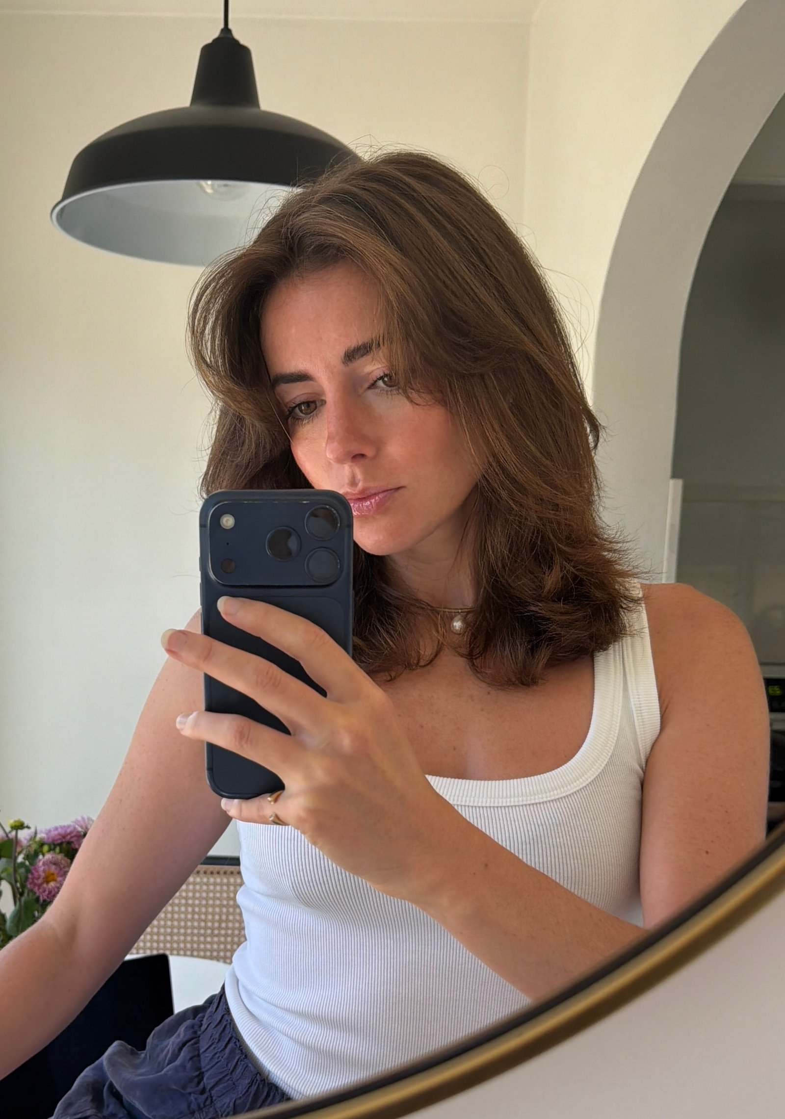 Woman with shoulder-length brown hair, wearing a white tank top, takes a mirror selfie with a smartphone in a well-lit room. A pendant light and some flowers are visible in the background.
