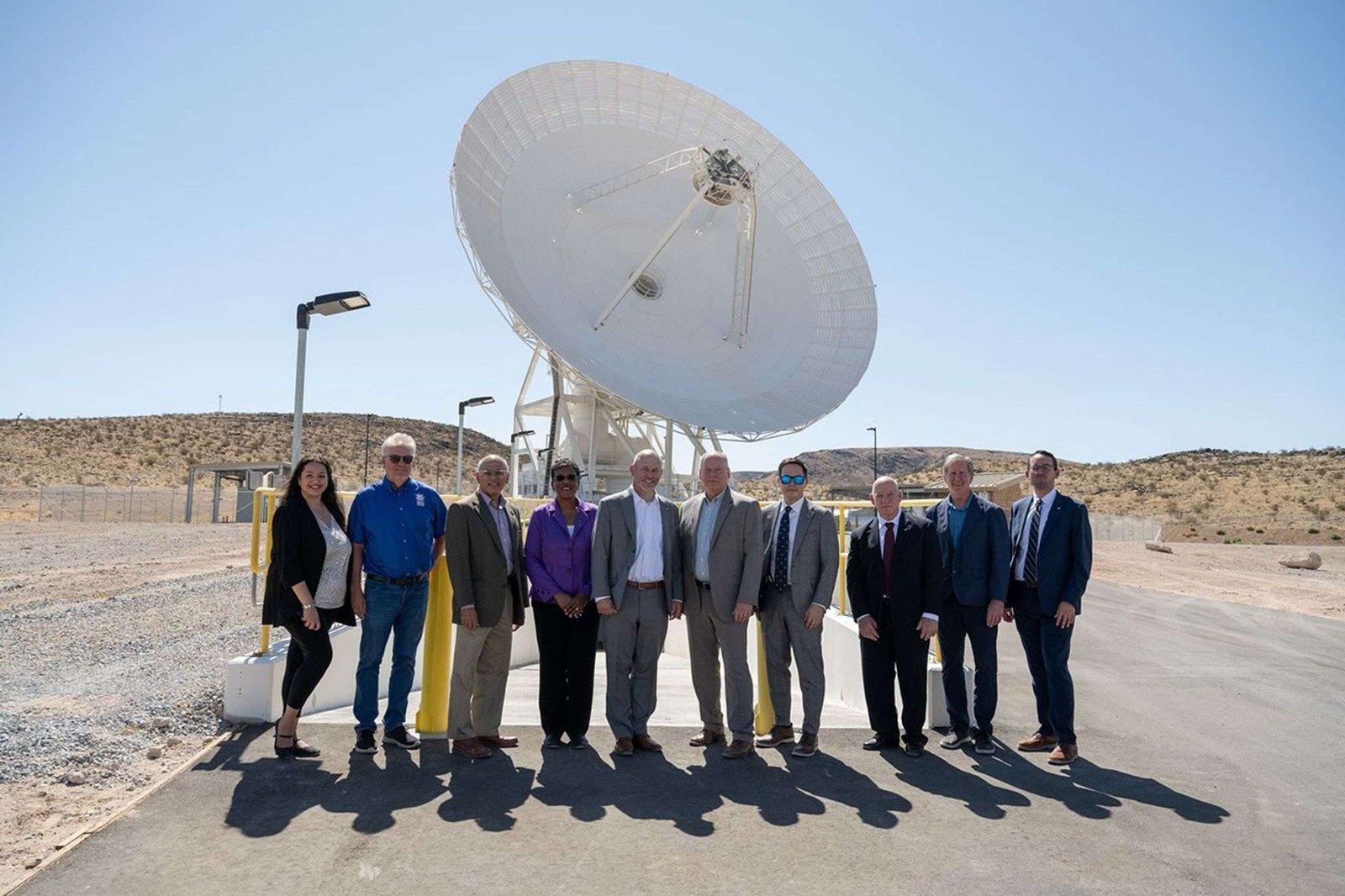 Ten people in professional attire pose together outside under a clear blue sky, with a massive white satellite dish standing directly behind them.