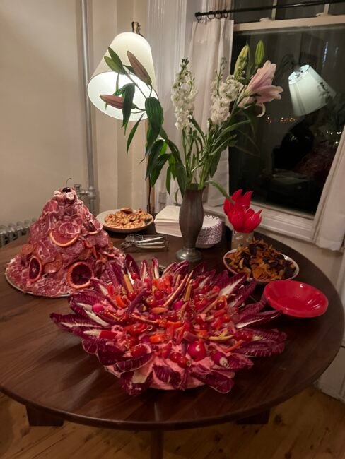 A table with a meat and radicchio platter, a meat-covered mound with citrus slices, small bowls of snacks, a vase of flowers, and a lamp, set in a dimly lit room.