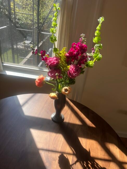 A vase of assorted flowers sits on a round wooden table in sunlight by a window, casting a shadow on the table.