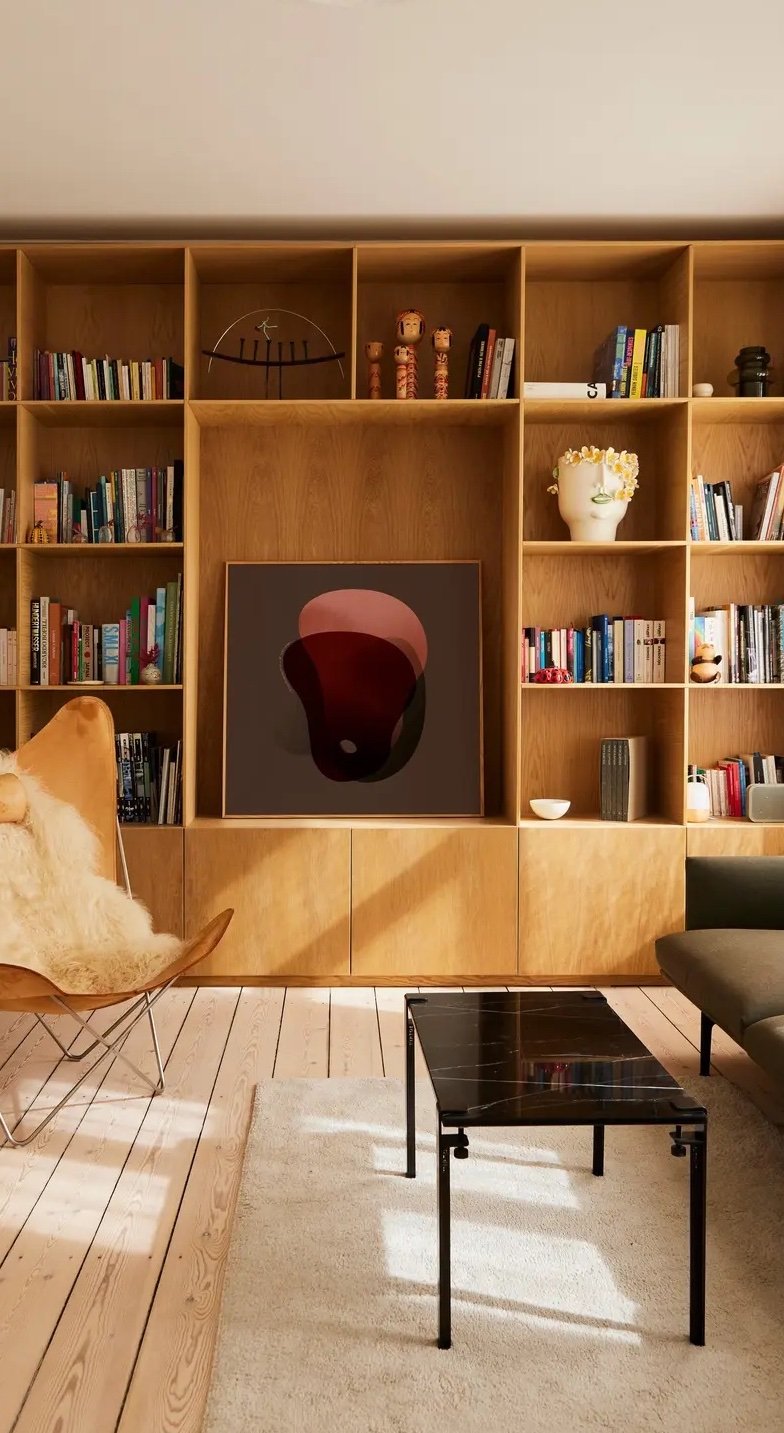 Modern living room with built-in wooden bookshelves, abstract art on a TV, a leather butterfly chair, black coffee table, and a white rug on light wood floors.
