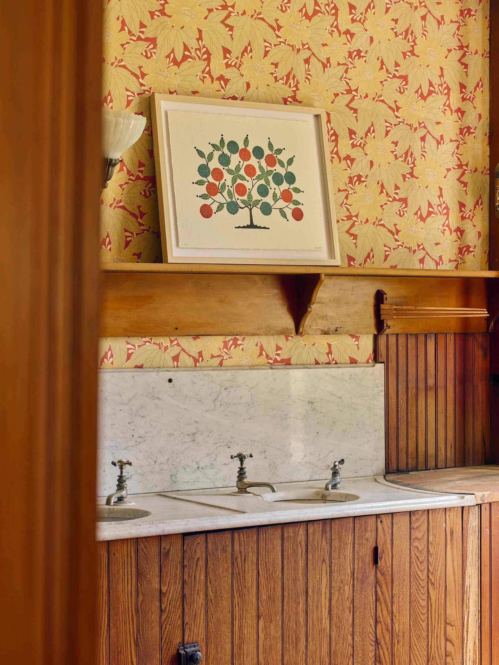 A framed tree illustration rests on a wooden shelf above a marble countertop with two faucets, set against floral-patterned wallpaper and wooden paneling.