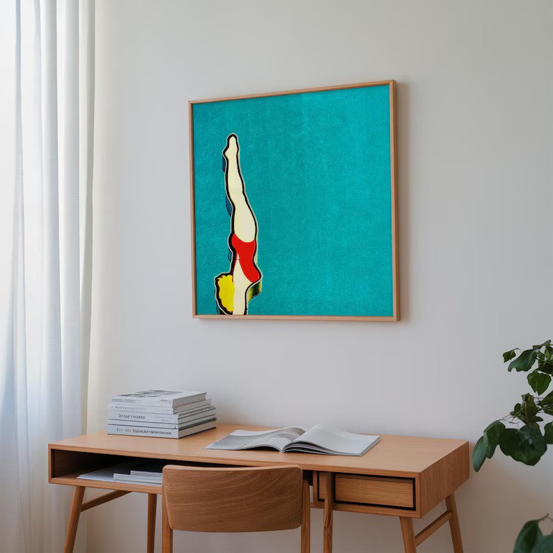 A desk with books and a plant sits beneath a framed artwork of a diver in a red swimsuit against a blue background hanging on a white wall.