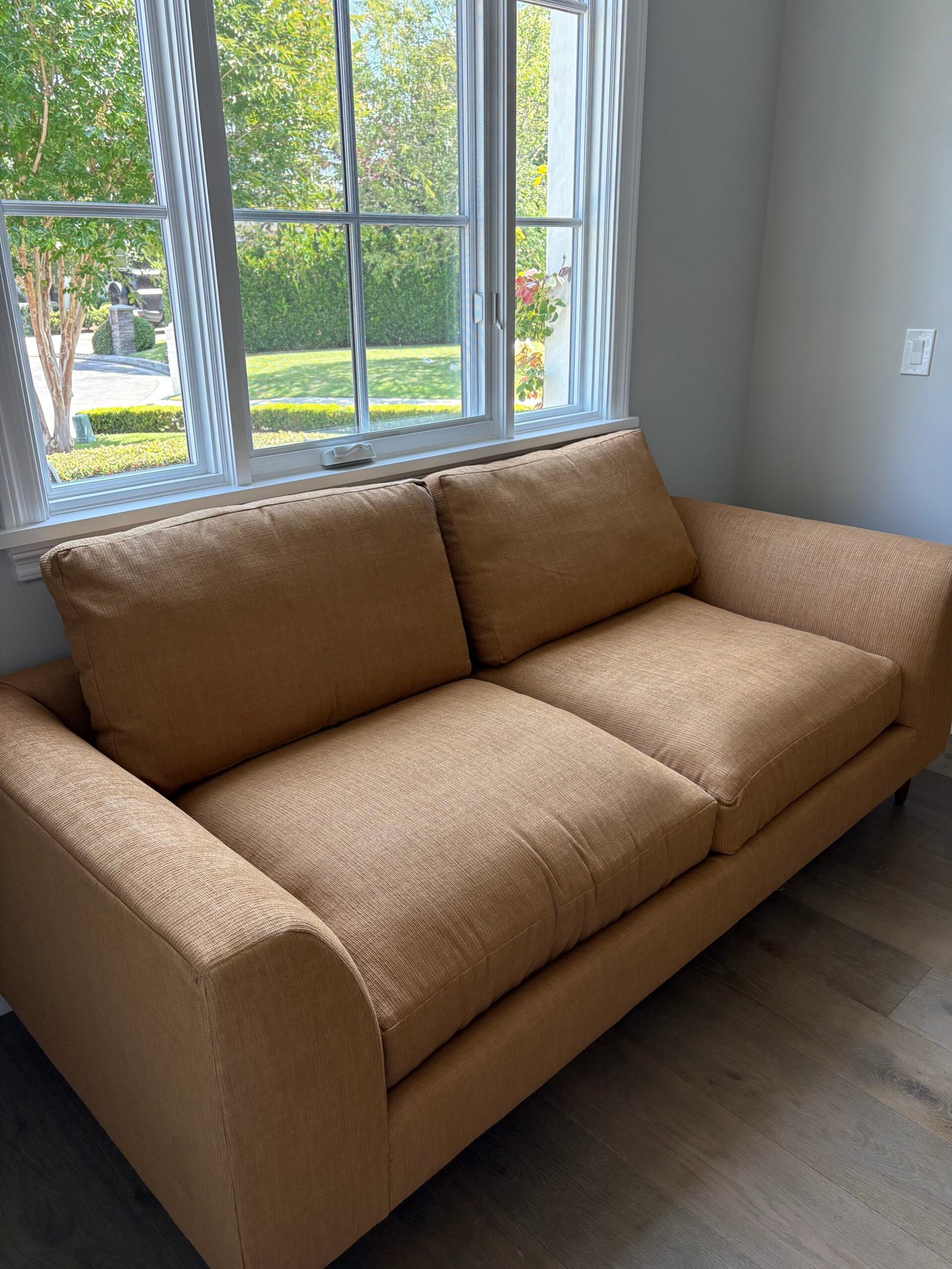 A tan fabric sofa with two seat cushions and two back cushions sits in front of a window with a view of greenery outside.