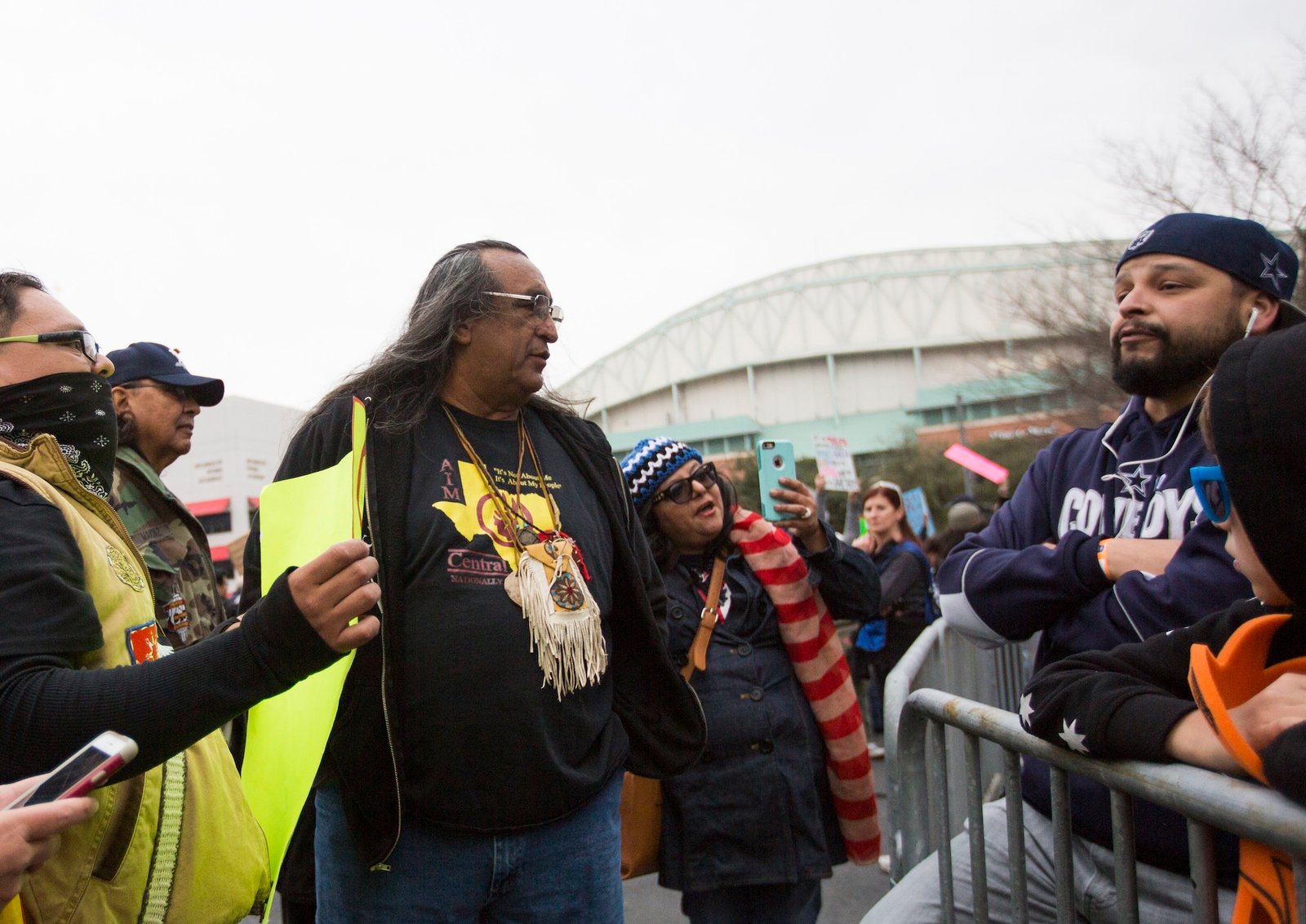 A man wearing a leather pouch around his neck talks to a person on the other side of a crowd barrier near a large building or stadium