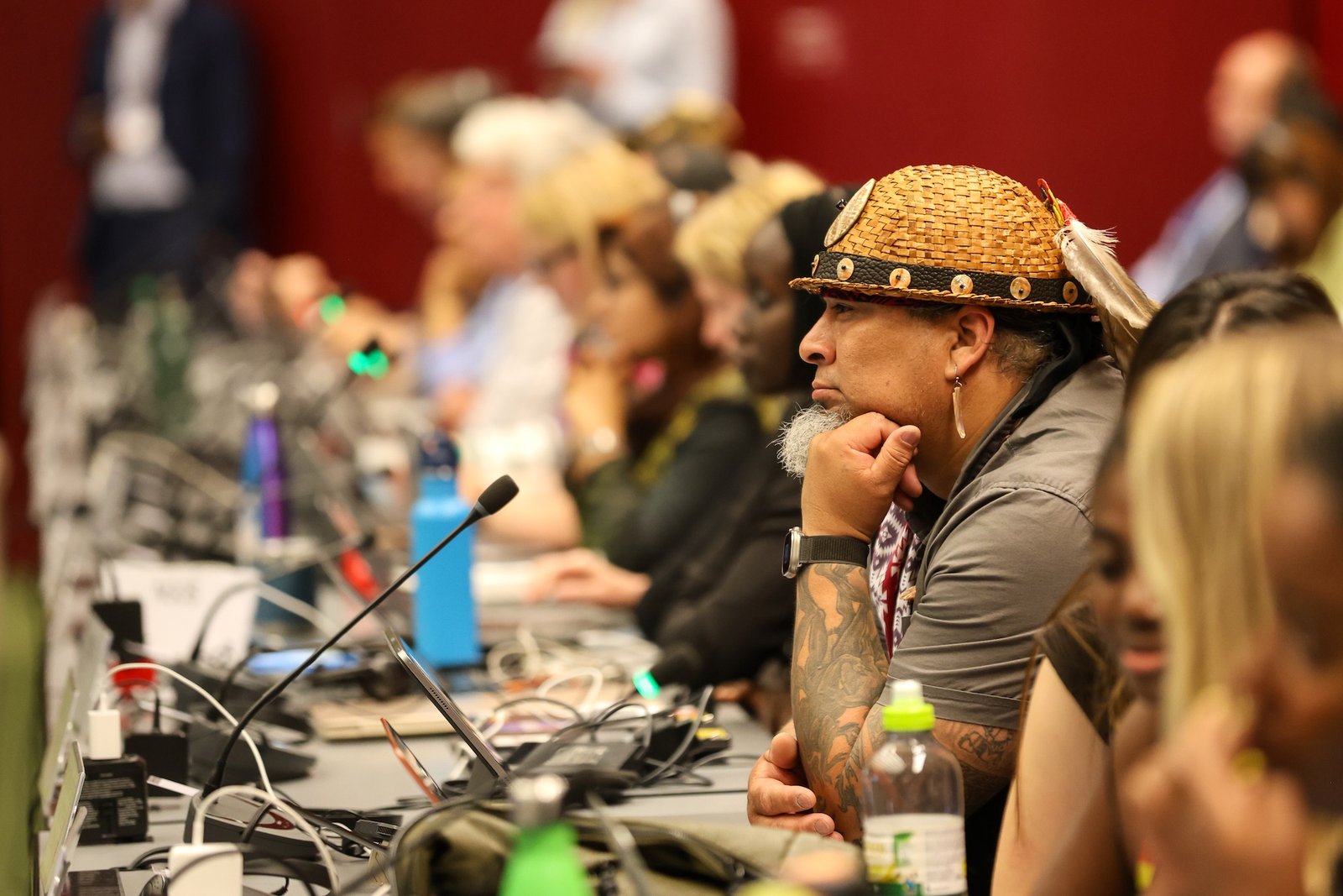 A man in a traditional hat sits on a long table with microphones, leaning forward as if listening