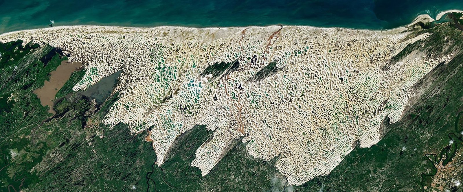 Bright white sand dunes stretch for dozens of kilometers between the dark blue Atlantic Ocean and green coastal vegetation. Blue and green freshwater lagoons fill low areas between the dunes, while dark-green vegetated patches lie within the dune field.