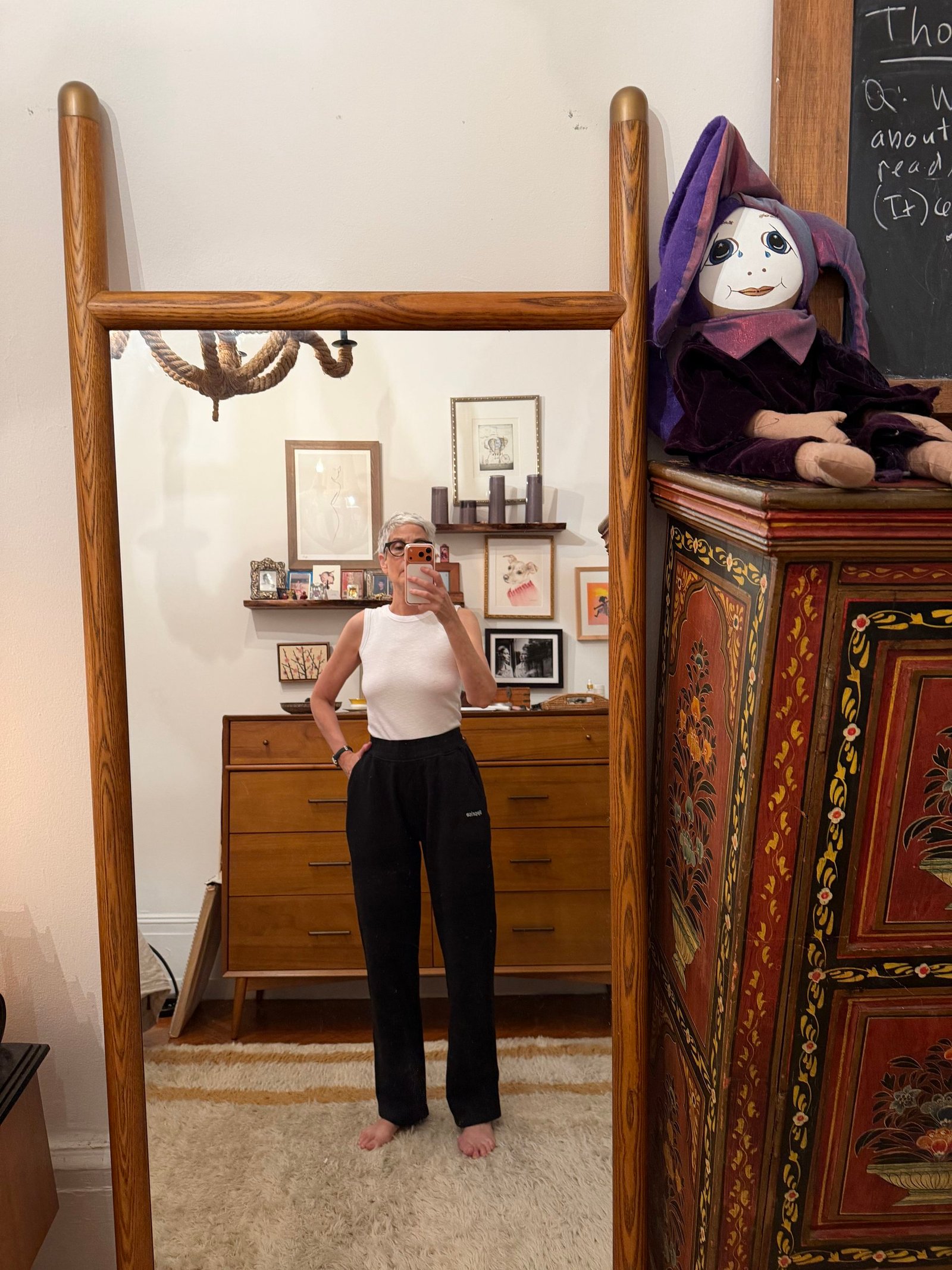 Person taking a mirror selfie in a room with mid-century furniture, framed pictures on the wall, and a decorative cabinet with a doll on top.
