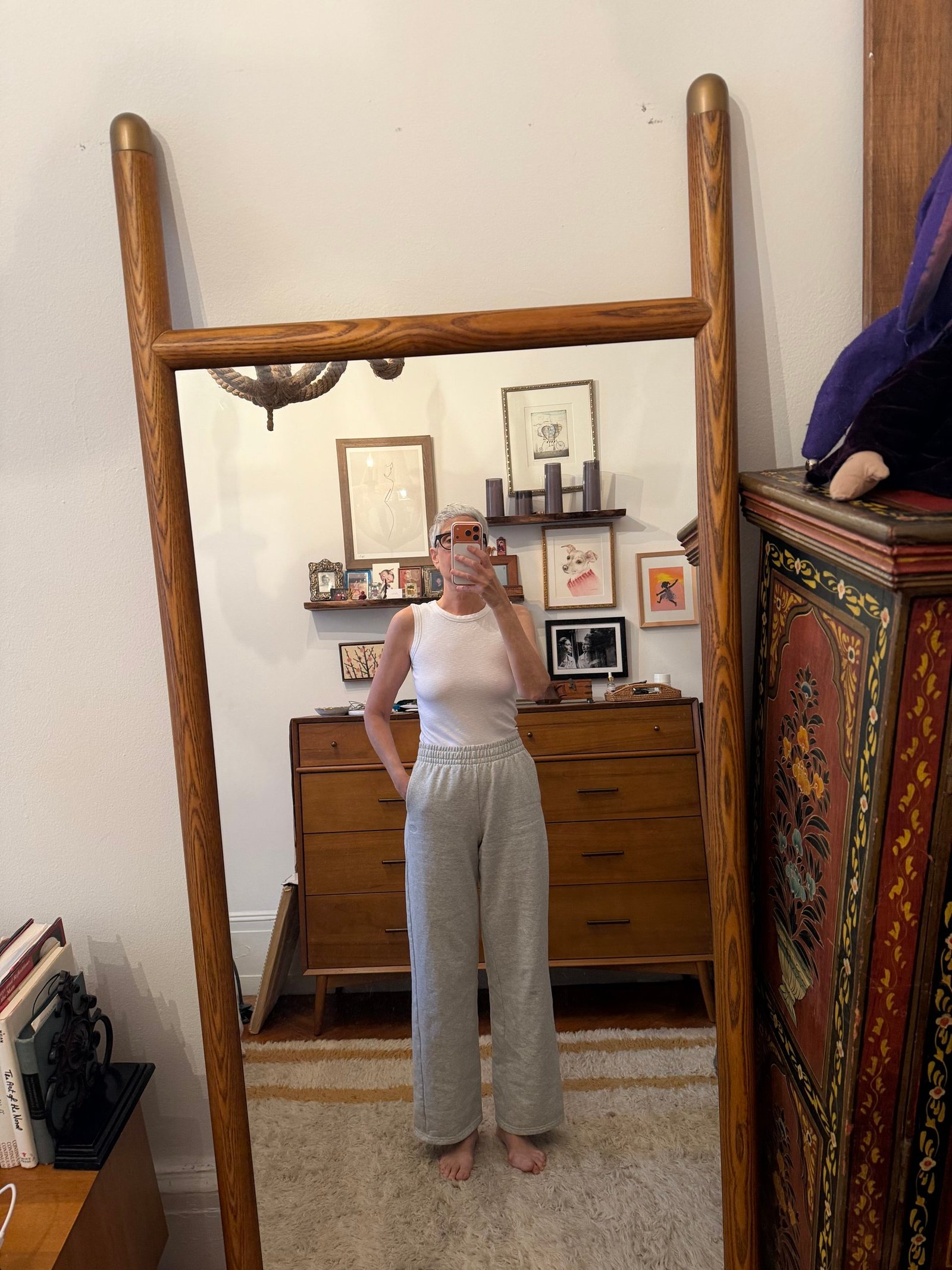 Person in a white sleeveless top and gray pants takes a mirror selfie in a room with wooden furniture, framed art, and books visible in the background.