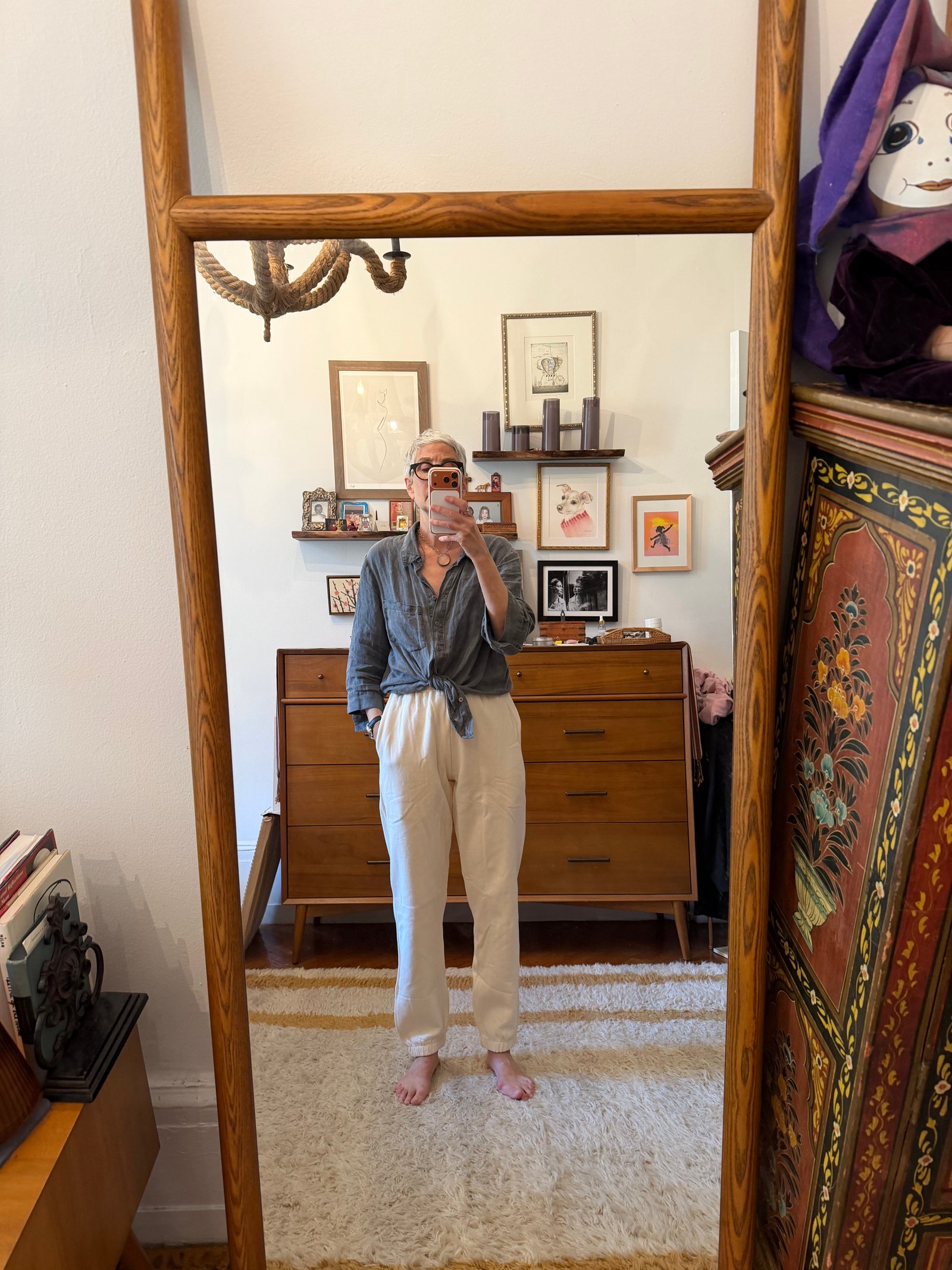 Person stands barefoot in front of a mirror, wearing a gray shirt and white pants. A wooden dresser, framed art, and a patterned cabinet appear in the background.