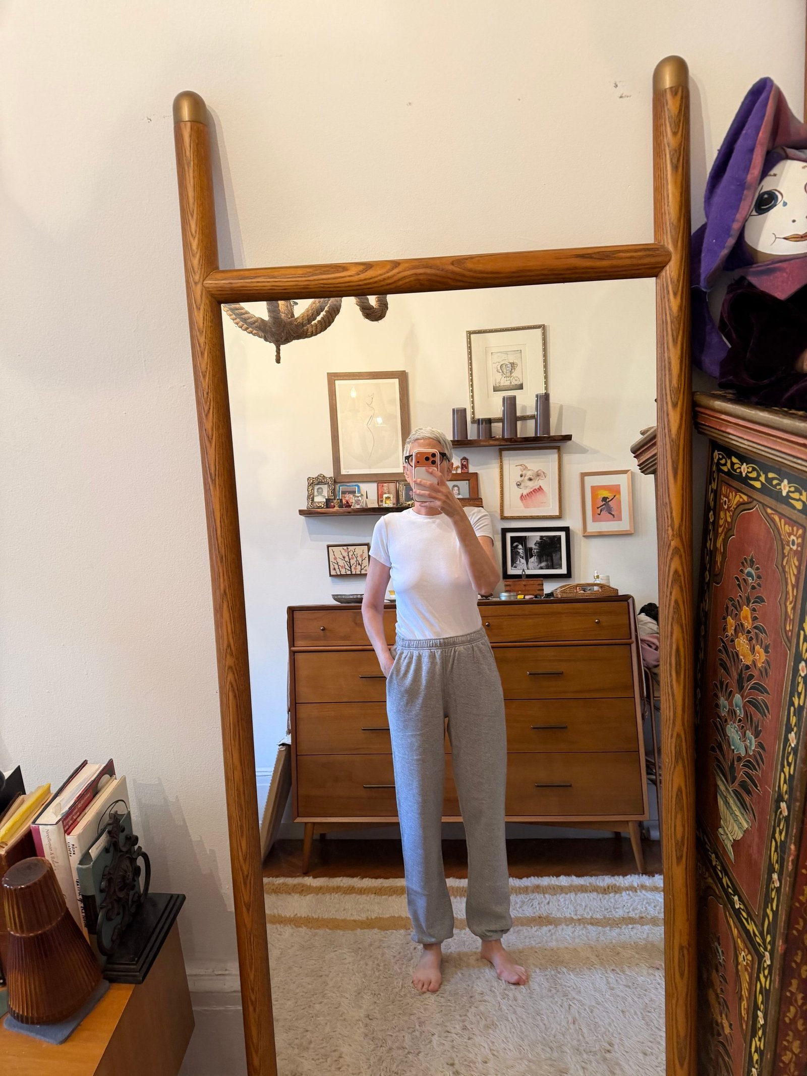 Person in a white shirt and gray sweatpants stands barefoot in front of a tall mirror in a room with wooden furniture and framed art on the wall.