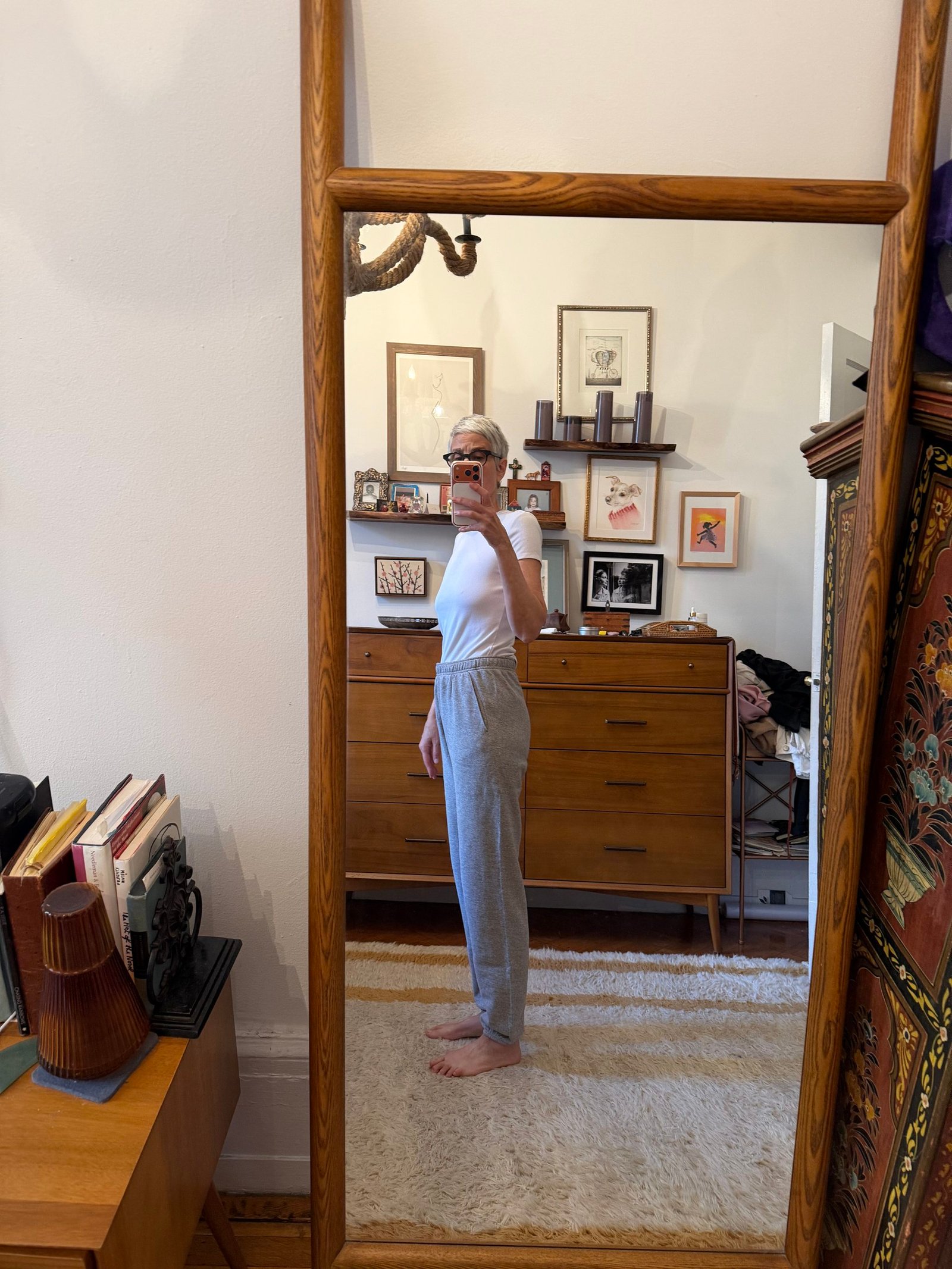 Person taking a side-view mirror selfie in a bedroom, wearing a white shirt and gray pants, with wooden furniture and framed art on the wall in the background.