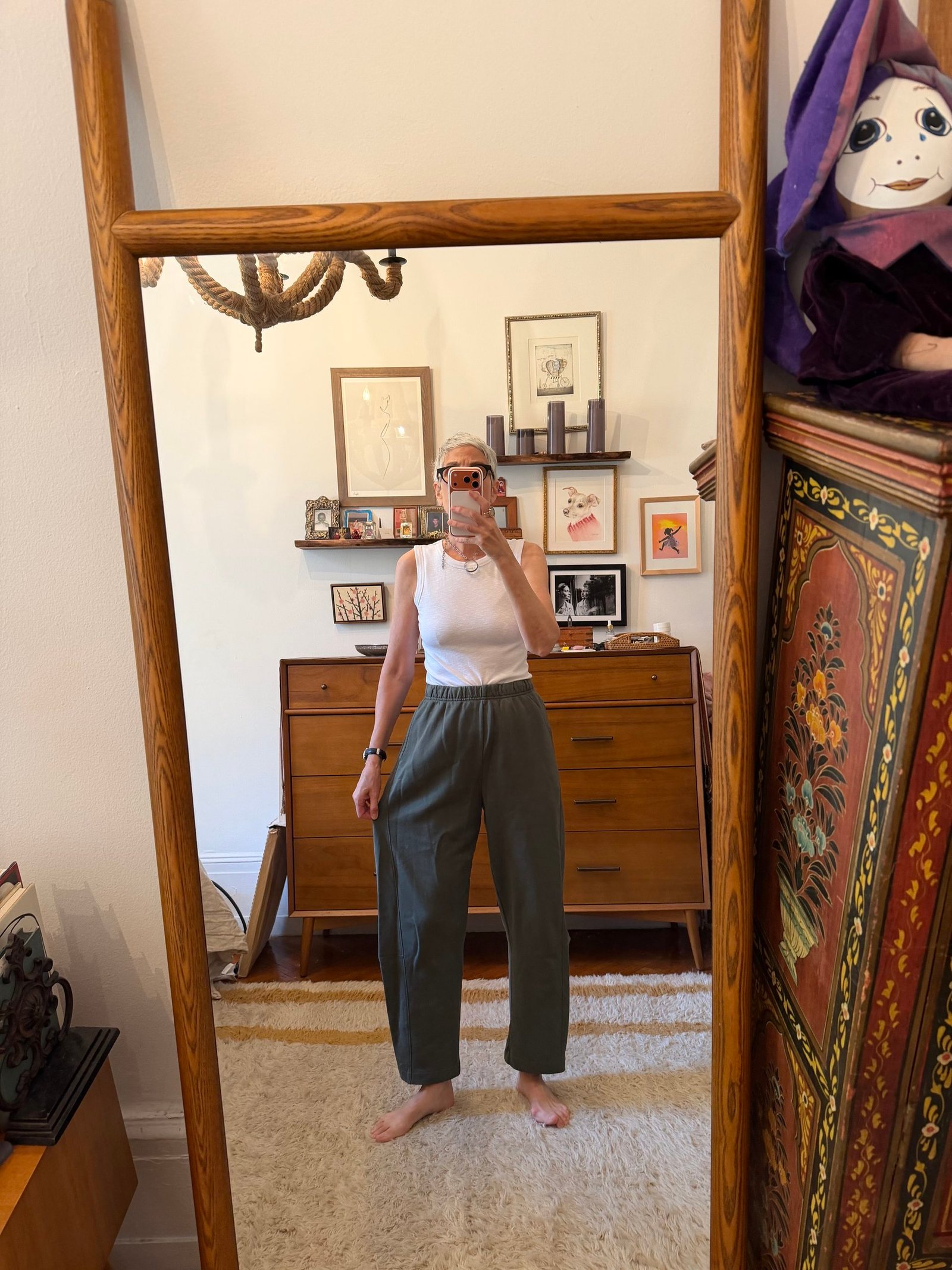 A person stands barefoot in front of a mirror, wearing a white sleeveless top and green pants, taking a selfie in a room with mid-century furniture and framed artwork on the wall.
