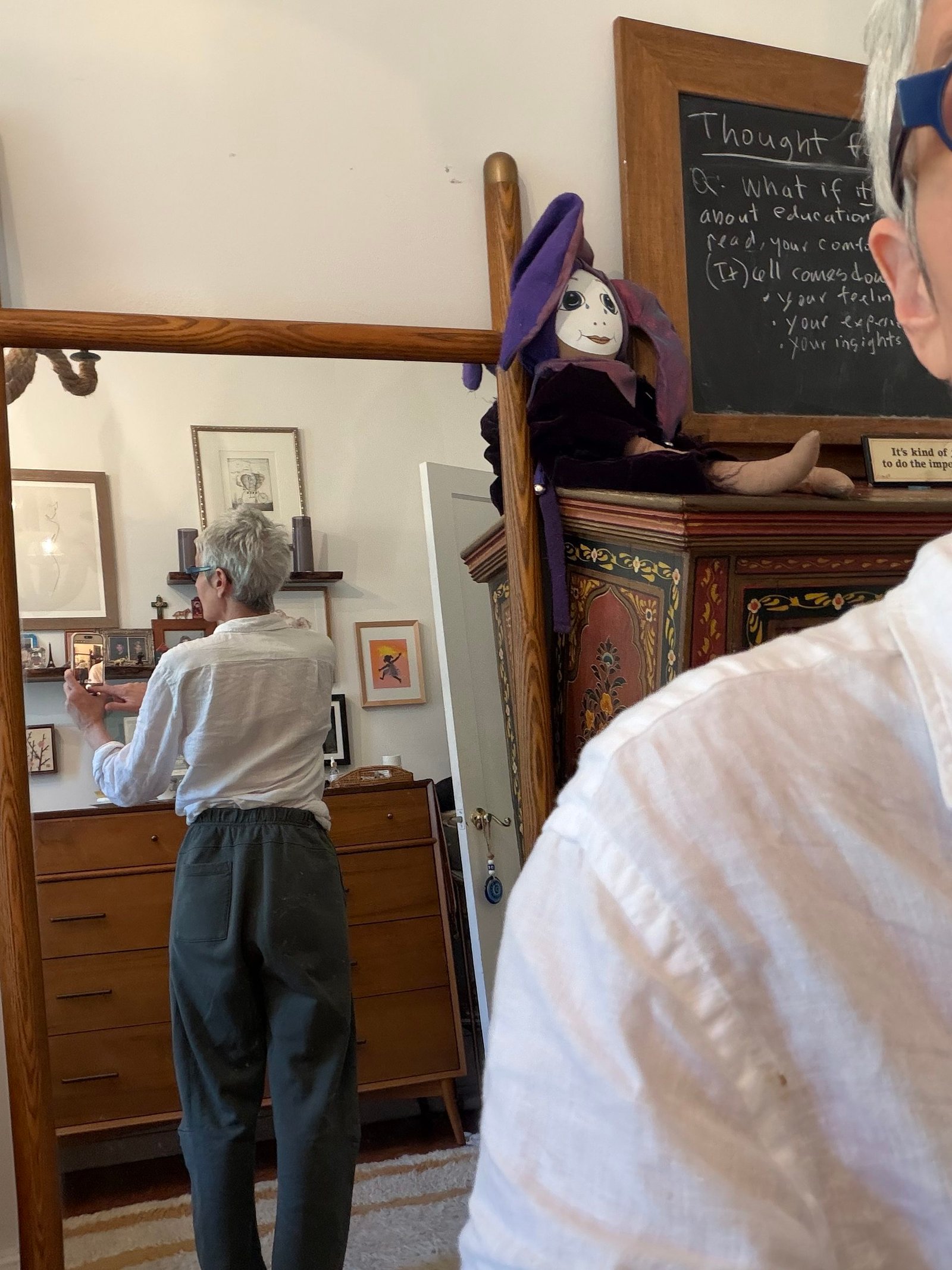 Person with short gray hair and glasses is seen from behind in a mirror, standing in a room with wooden furniture, framed pictures, and a purple-hat doll on top of a cabinet.