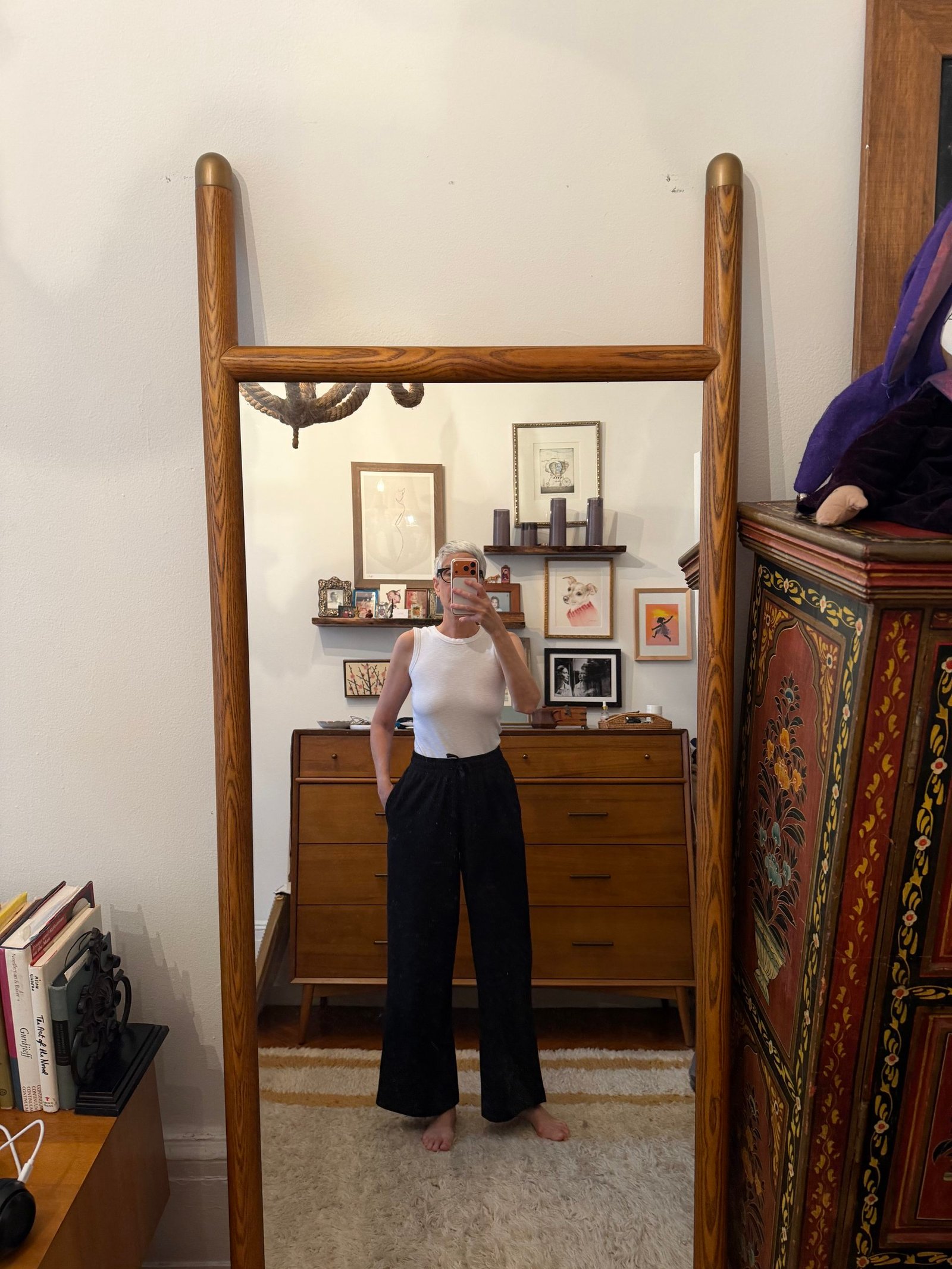 Person in a white sleeveless top and black pants takes a mirror selfie in a mid-century style room with framed art and wooden furniture.