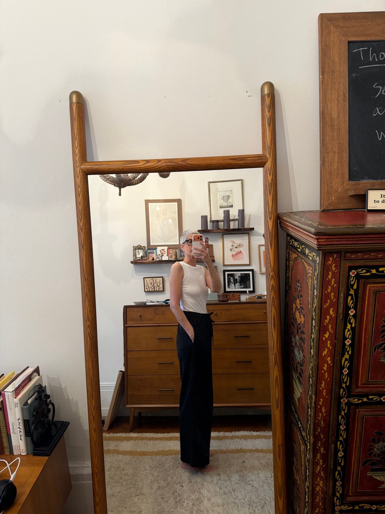 Person in white sleeveless top and dark pants takes a mirror selfie in a room with wooden furniture, framed artwork, books, and a decorative cabinet.
