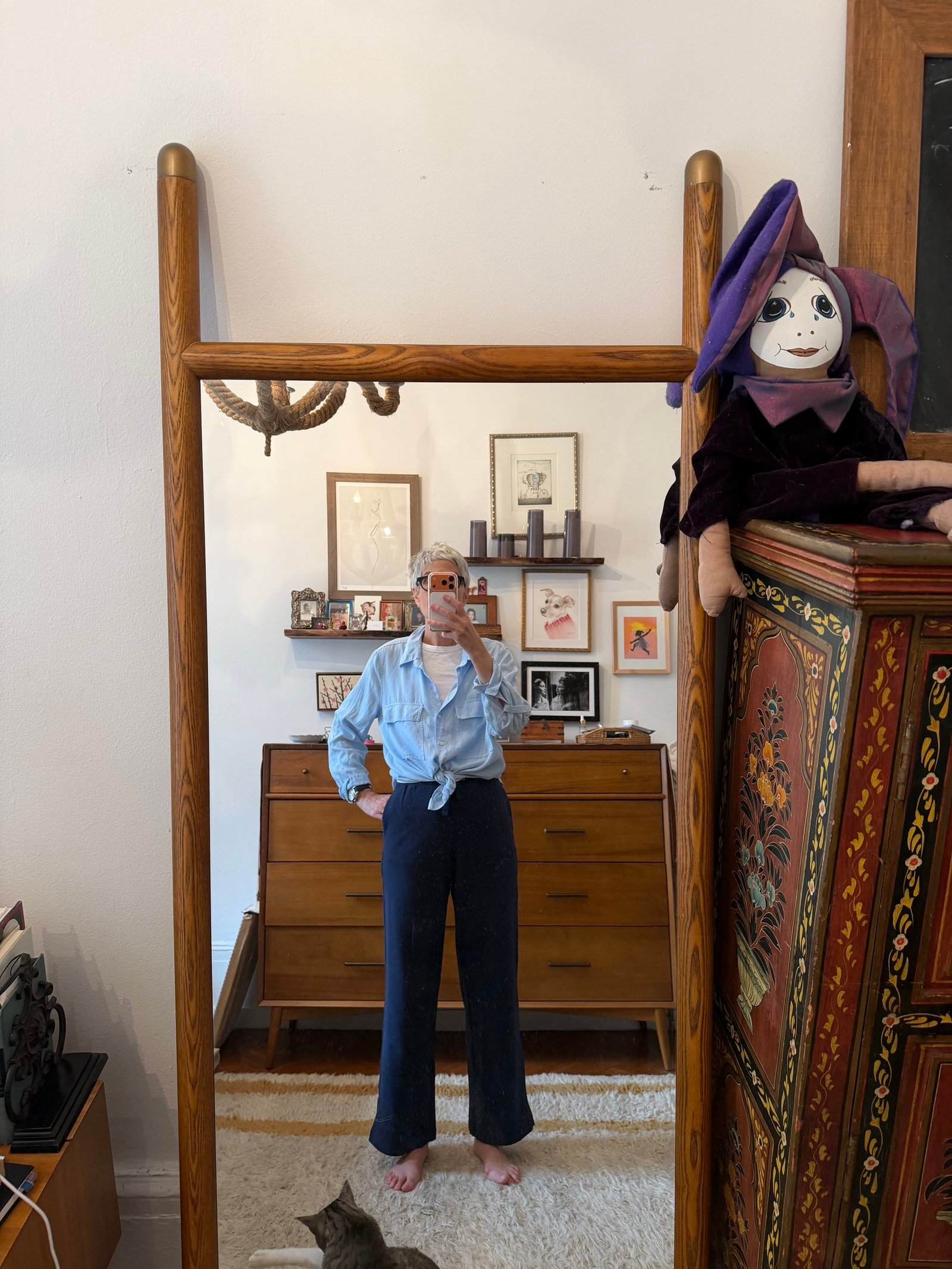 Person taking a mirror selfie in a room with mid-century furniture; a large doll sits on a cabinet, and a cat is visible on the floor.