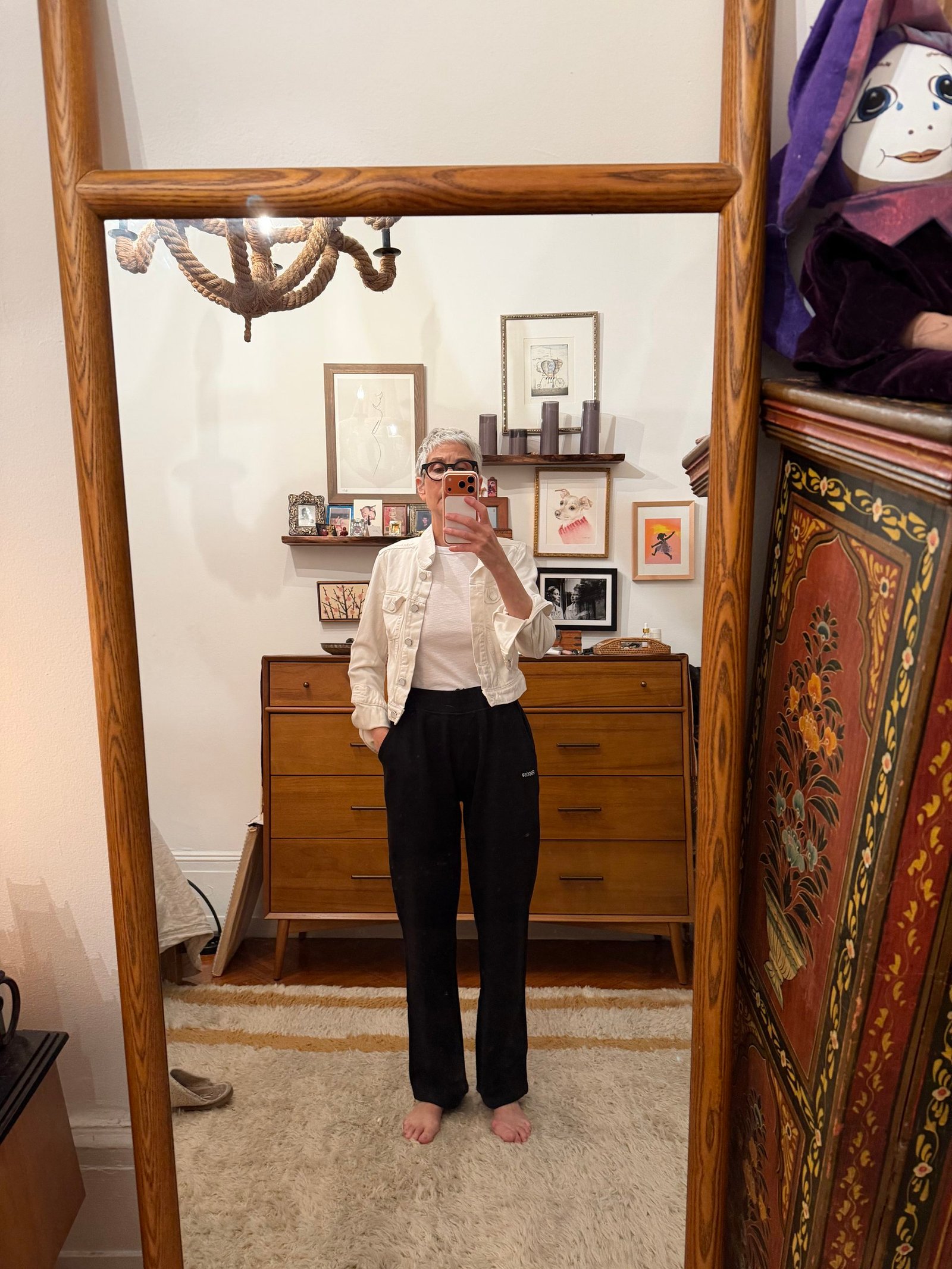 Person stands barefoot in front of a mirror, wearing black pants, a white shirt, and sunglasses. The room has wooden furniture, framed art on the wall, and a decorative cabinet on the right.