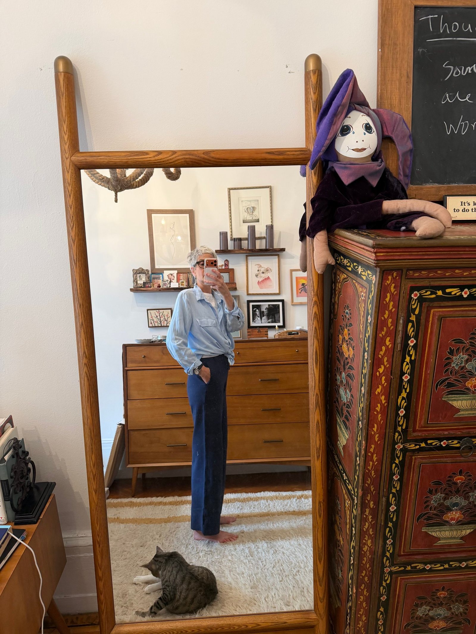 Person taking a mirror selfie in a bedroom with a cat lying on the rug and a doll in a purple outfit sitting on a decorated cabinet.