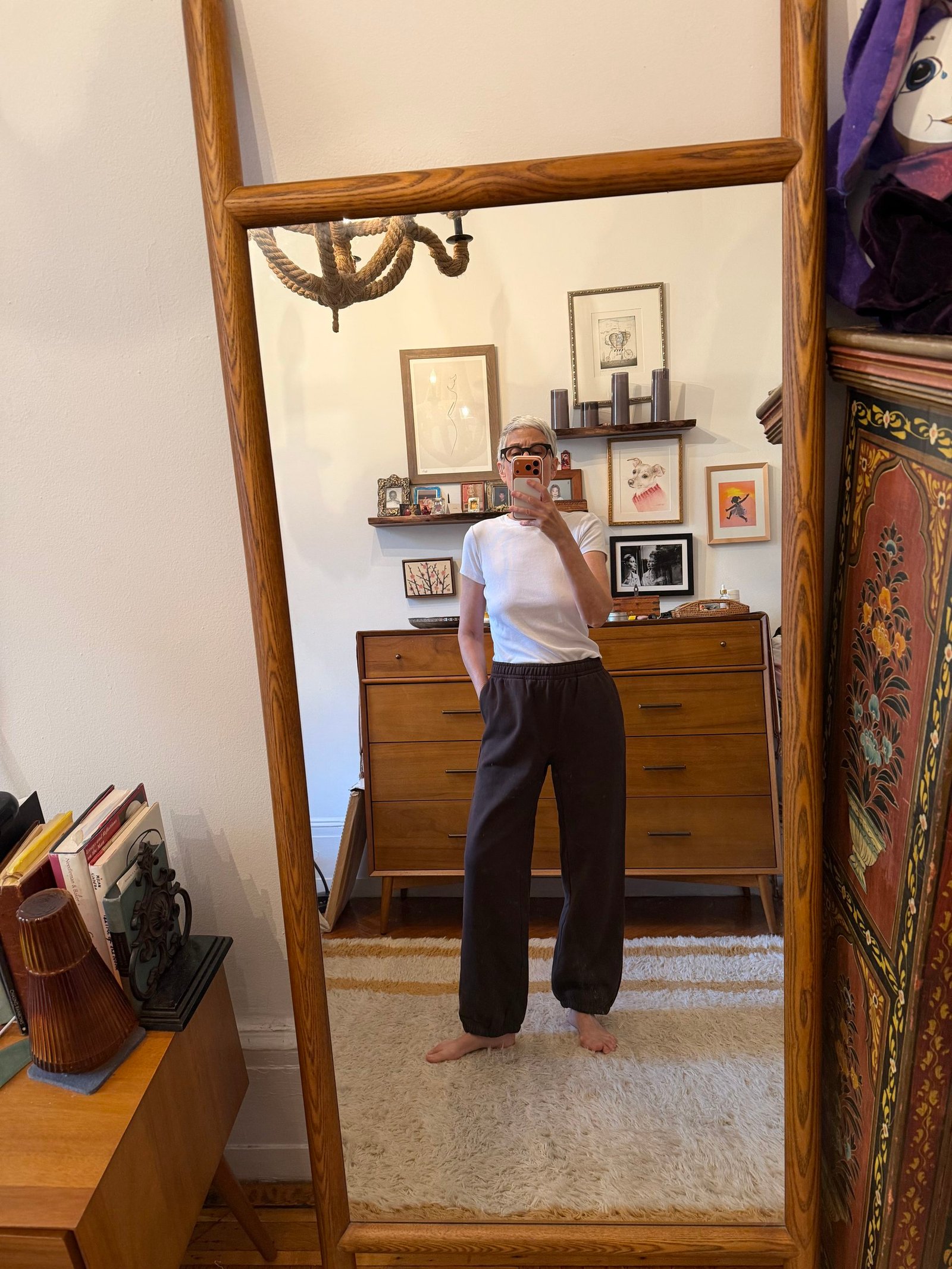 Person takes a mirror selfie in a room with mid-century furniture, art on the walls, and books on a shelf. They wear a white sleeveless top and dark pants, standing barefoot on a rug.
