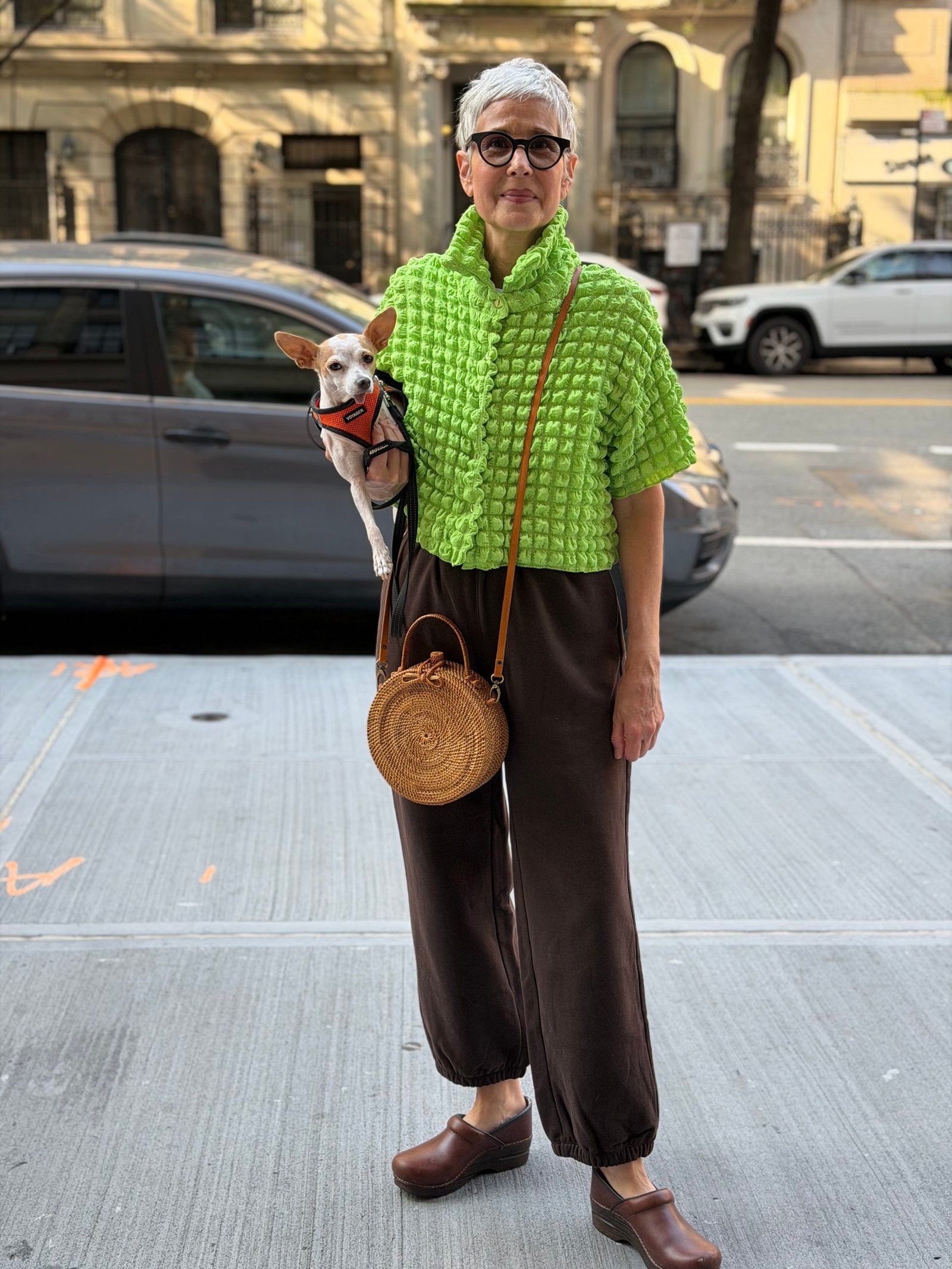 Woman with short gray hair wearing glasses, a bright green textured top, brown pants, and brown clogs stands on a city sidewalk holding a small dog and a round straw bag.