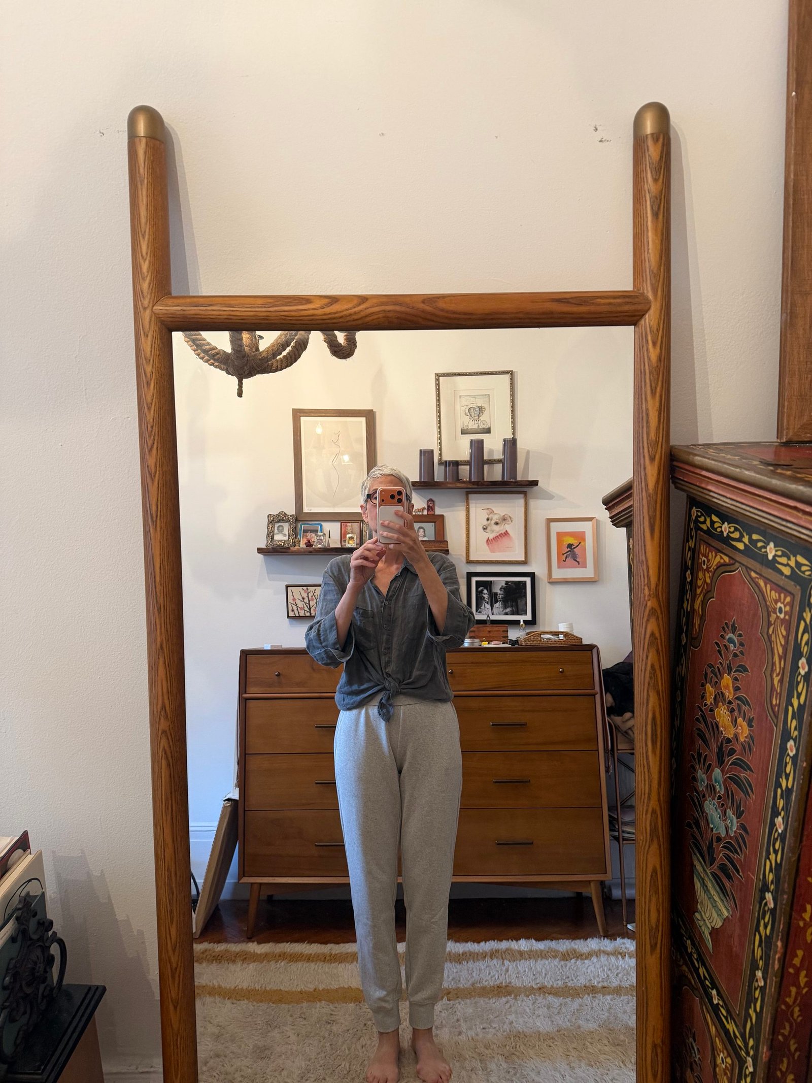 Person taking a mirror selfie in a cozy room with wooden furniture, framed art on the wall, and a patterned cabinet to the right.