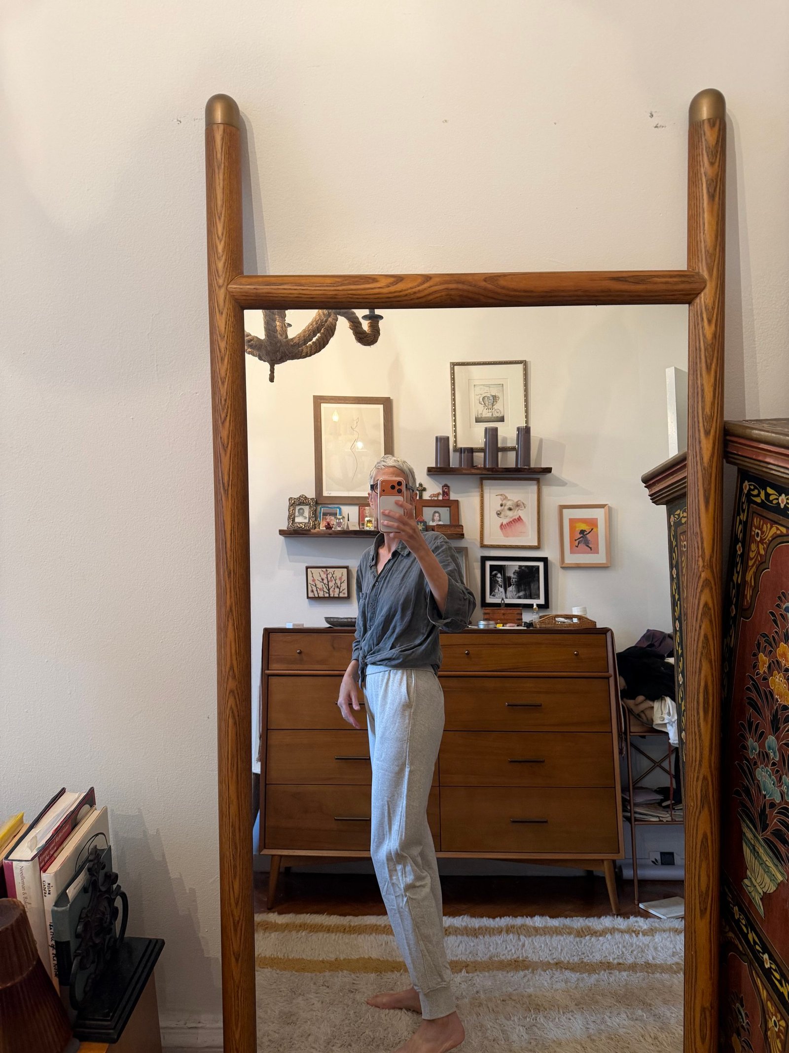 Person taking a mirror selfie in a room with wooden furniture, art on the walls, and books on the floor; wearing a gray shirt and light gray pants.