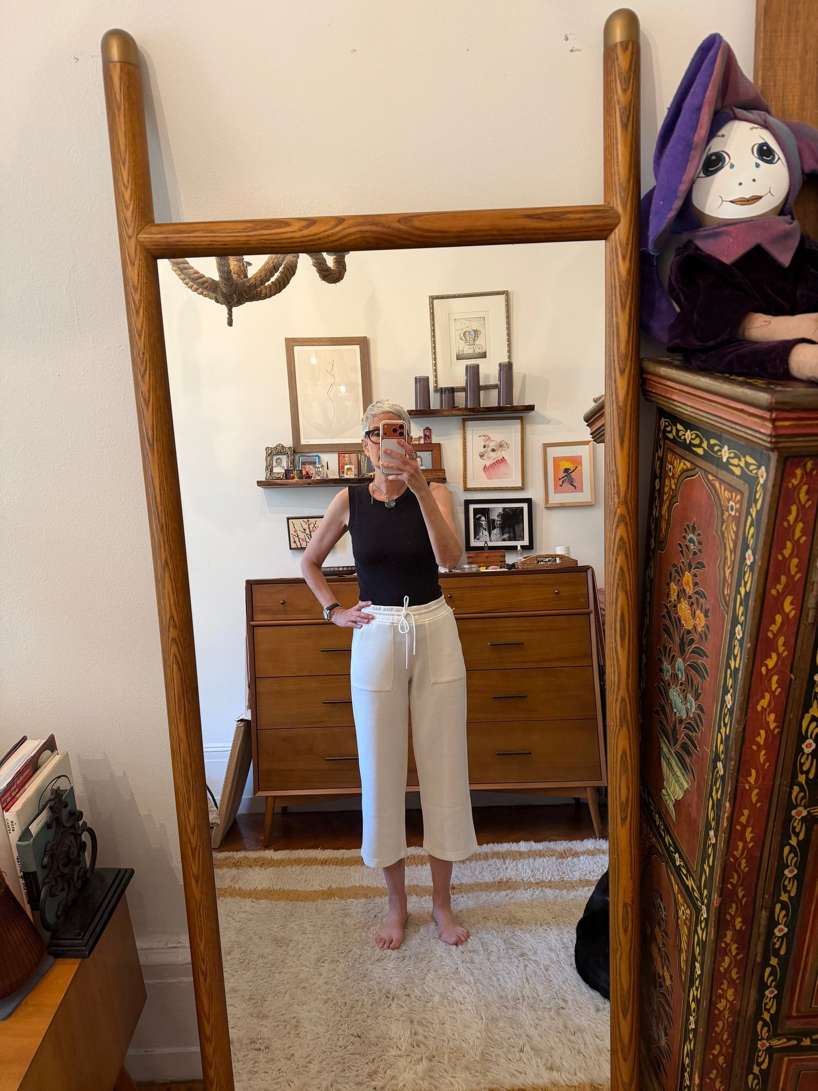 Person takes a mirror selfie in a room with eclectic decor, wearing a black sleeveless top and white pants. Various framed pictures and a large purple puppet are visible in the background.
