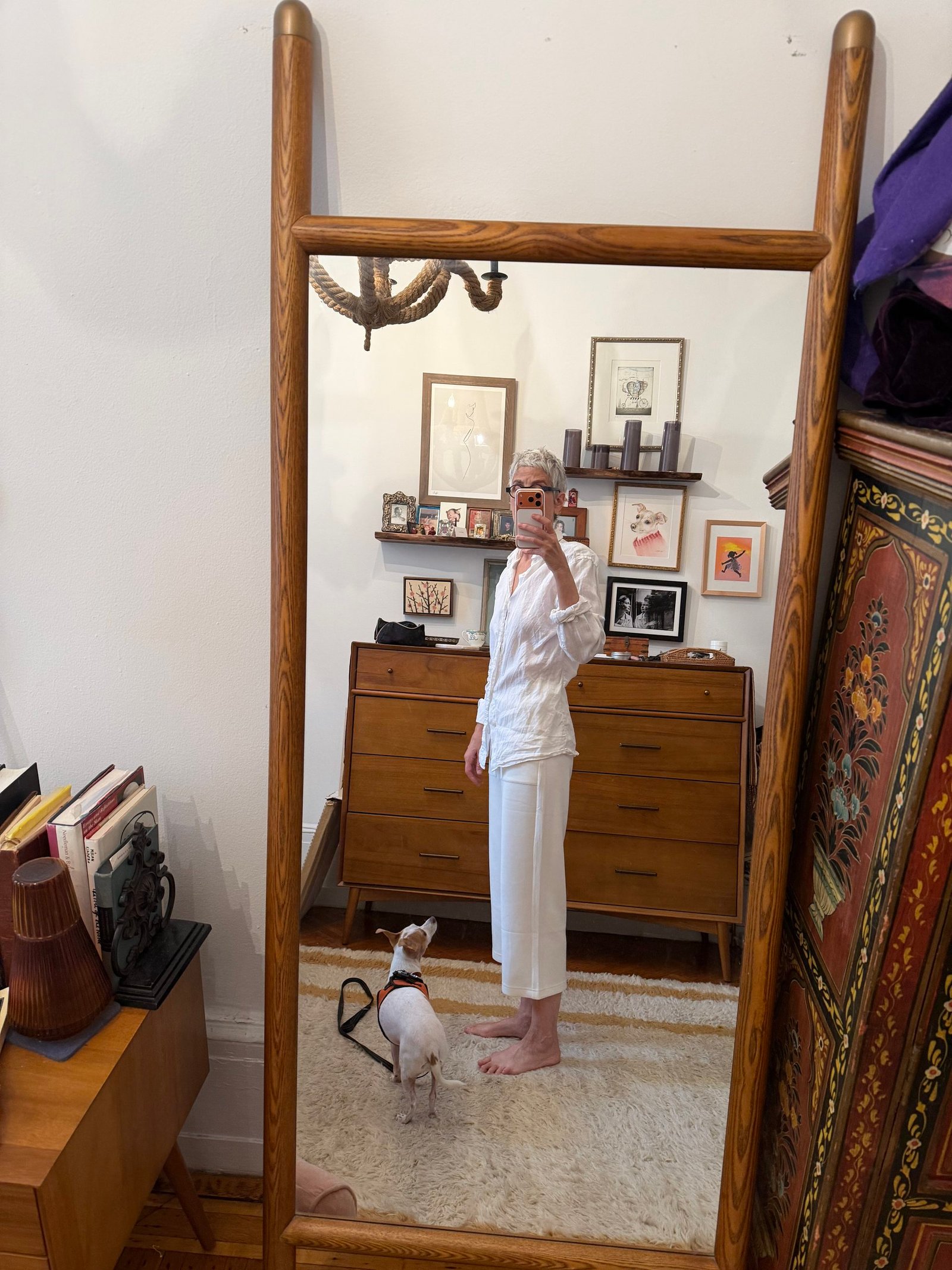 Person in white clothes taking a mirror selfie in a room with a small dog, wooden furniture, art on the wall, and various items on surfaces.