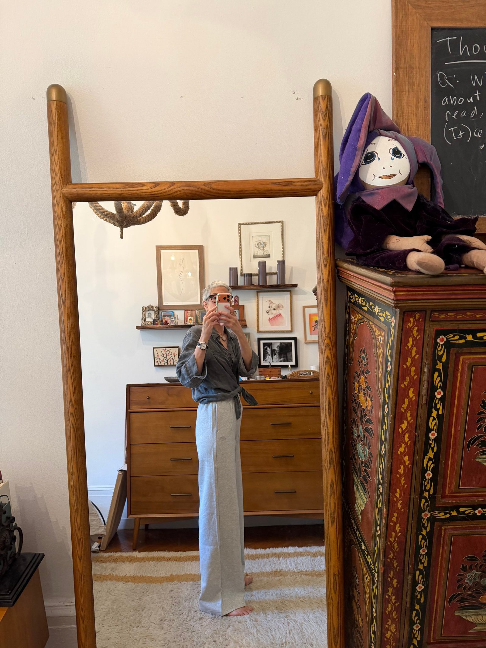 Person taking a mirror selfie in a room with wooden furniture, wall art, a rug, and a doll sitting on a decorated chest.