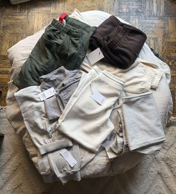 Assorted sweatpants and sweatshirts in neutral colors are arranged in a pile on a white cushion, each with brand tags attached, on a wooden floor.