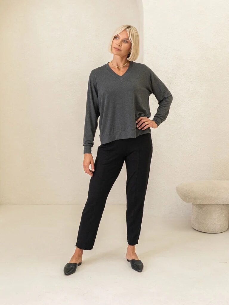 Woman standing indoors wearing a grey long-sleeve top, black pants, and black shoes, posing with one hand on her hip against a minimalist white background with a bench.