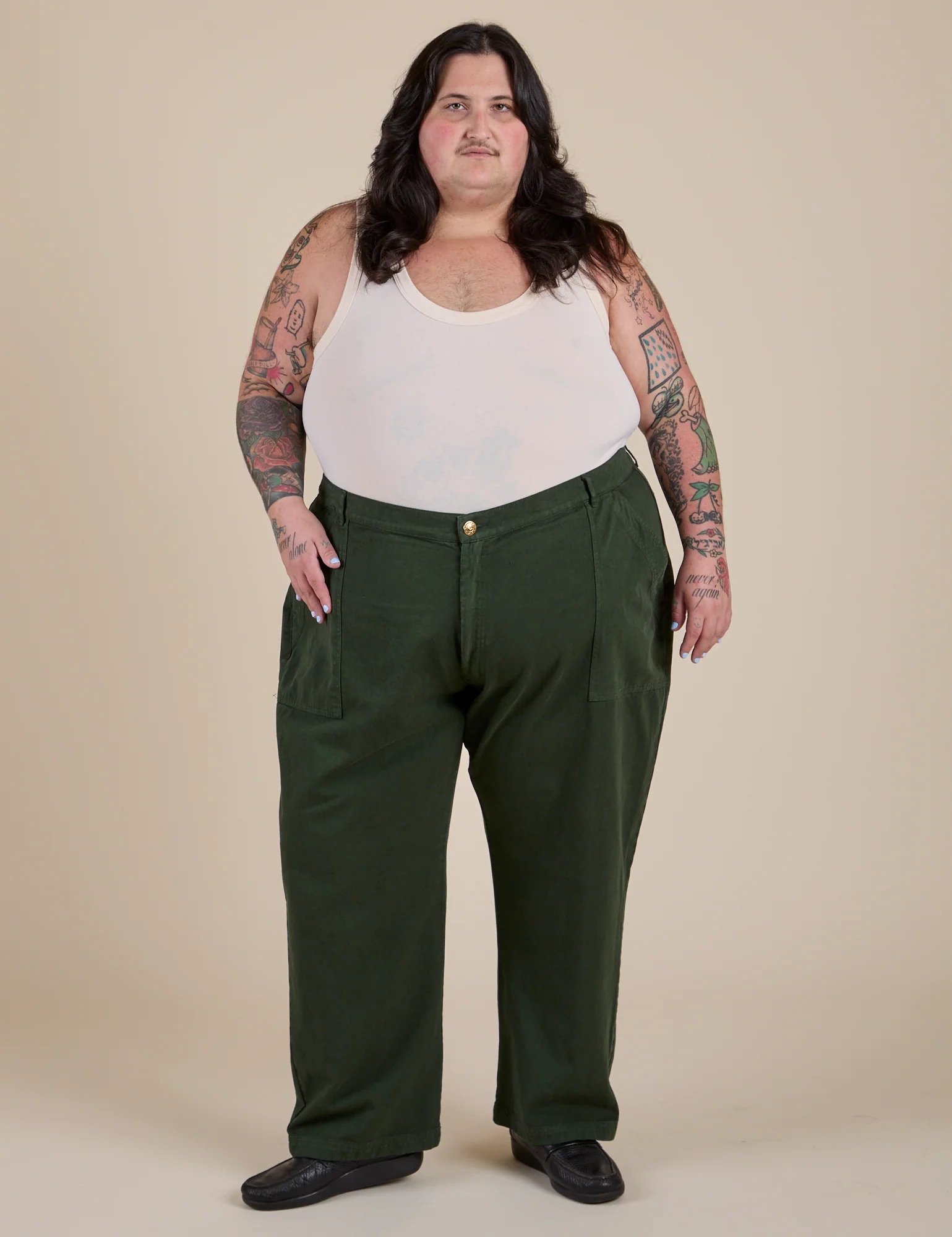 Person with long dark hair, tattoos, white tank top, and green pants stands facing forward against a plain beige background.
