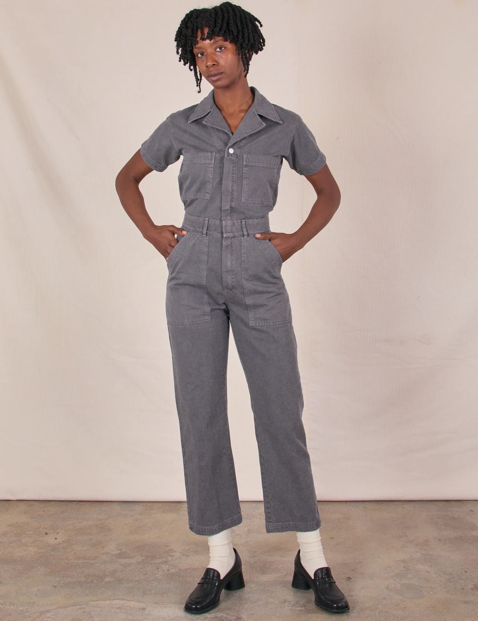 Person standing indoors wearing a gray short-sleeve jumpsuit, white socks, and black loafers, with hands on hips against a neutral backdrop.
