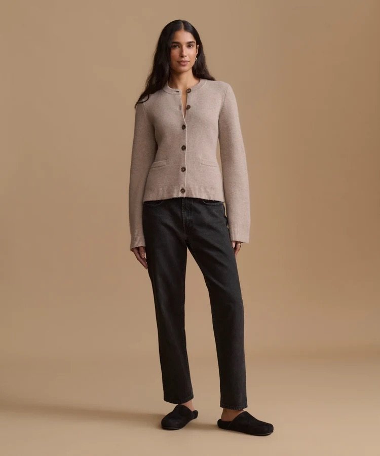 A person stands facing forward, wearing a beige button-up cardigan, black pants, and black slippers against a plain beige background.