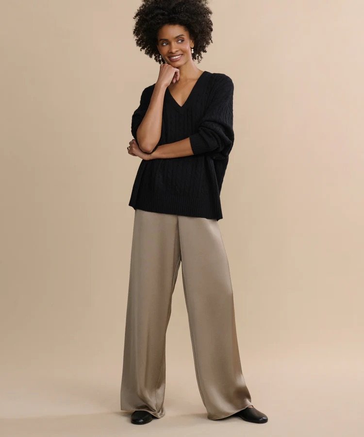 A person stands against a beige background, wearing a black V-neck sweater, light brown wide-leg pants, and black shoes, smiling with one hand touching their face.