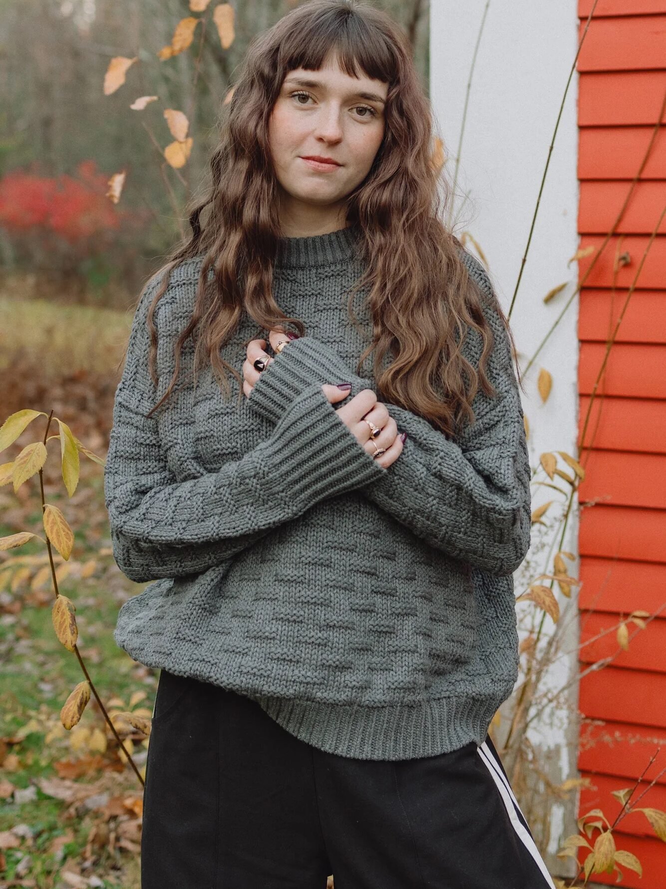 Person with long brown wavy hair wearing a gray knit sweater and black pants stands outside near an orange building, surrounded by autumn foliage.