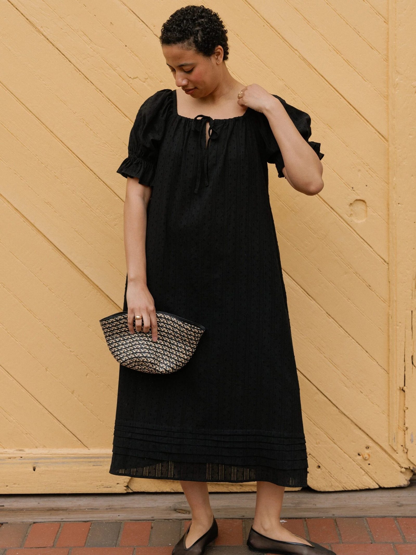 Person in a black, short-sleeved, ankle-length dress holds a woven clutch and looks down, standing against a light yellow wooden wall.