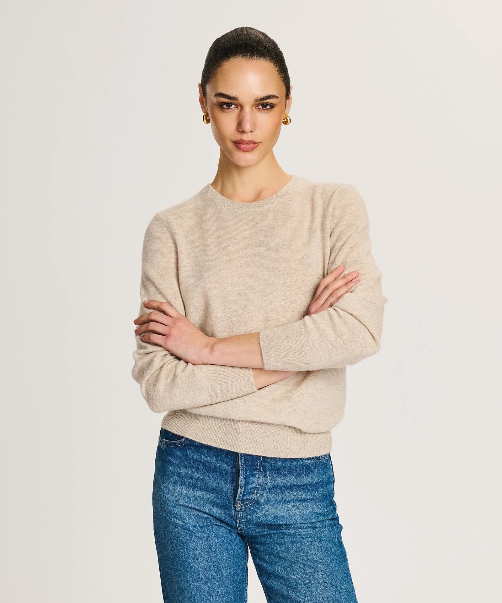A woman stands against a plain background, wearing a beige sweater and blue jeans, with her arms crossed and a neutral facial expression.