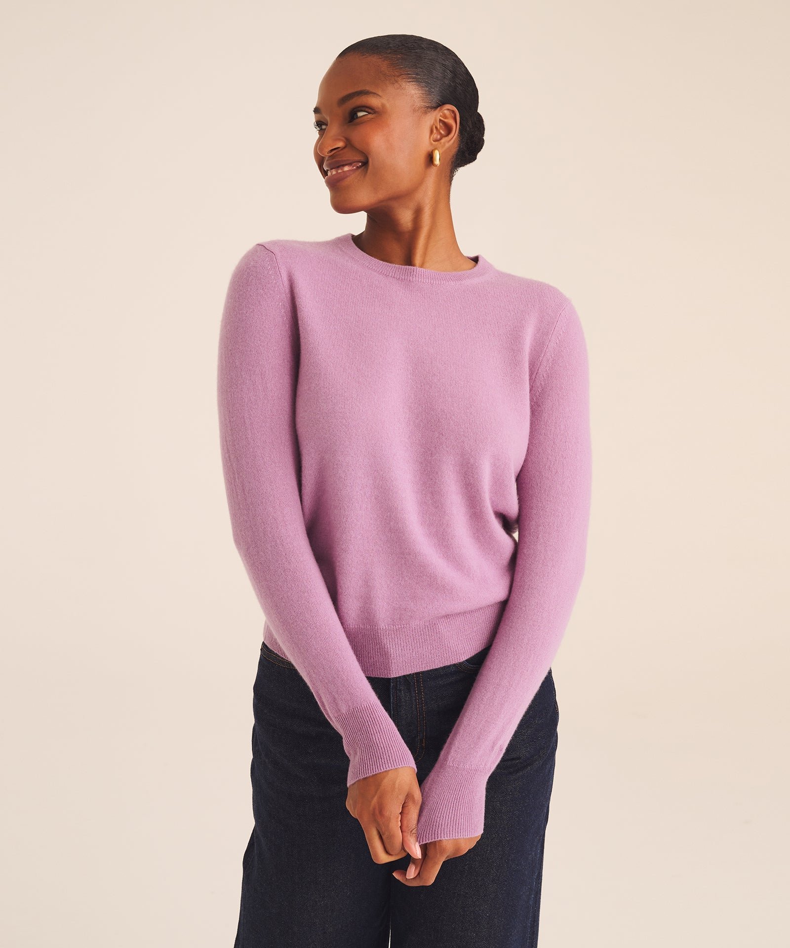 A person stands smiling, wearing a light purple long-sleeve sweater and dark jeans against a plain beige background.