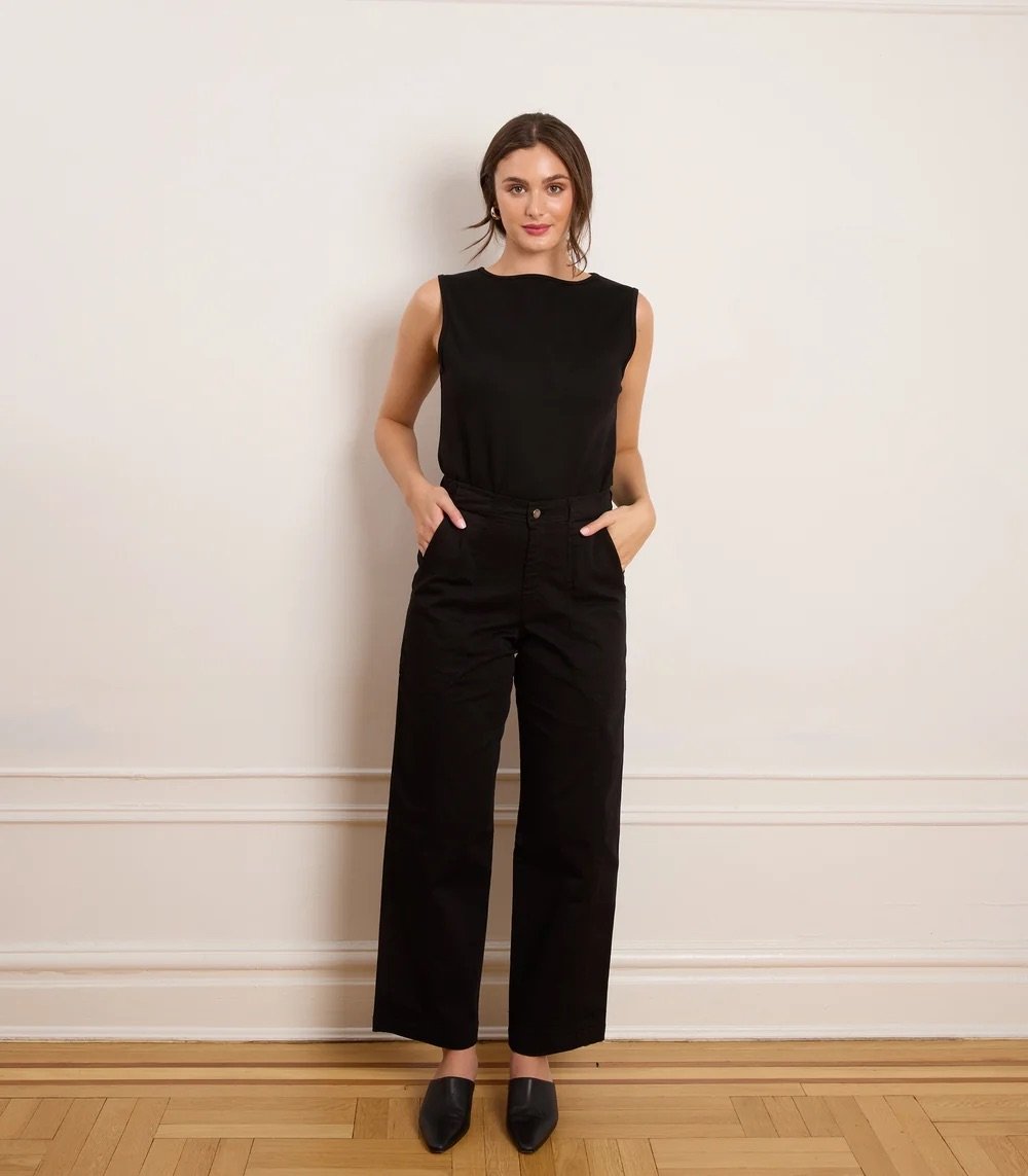 Woman stands indoors against a white wall, wearing a sleeveless black top, high-waisted black pants, and black shoes, with hands in her pockets.