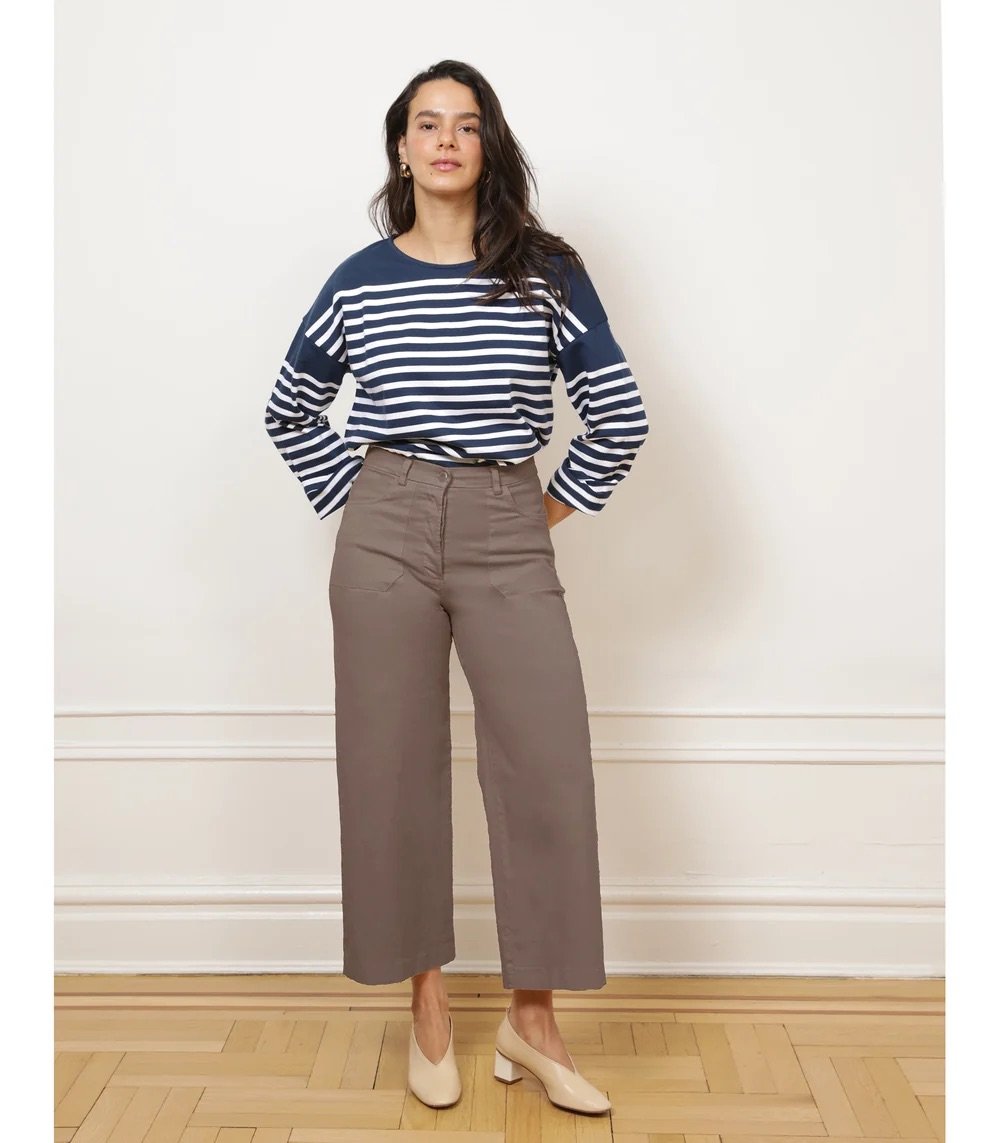 A woman stands indoors wearing a navy and white striped long-sleeve shirt, taupe high-waisted wide-leg pants, and beige heels against a plain wall and wooden floor.