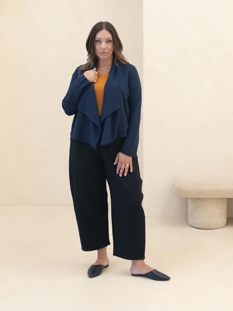 Woman standing indoors wearing a navy blue cardigan, mustard yellow top, black wide-leg pants, and black slip-on shoes, with a neutral background and a minimalist bench.