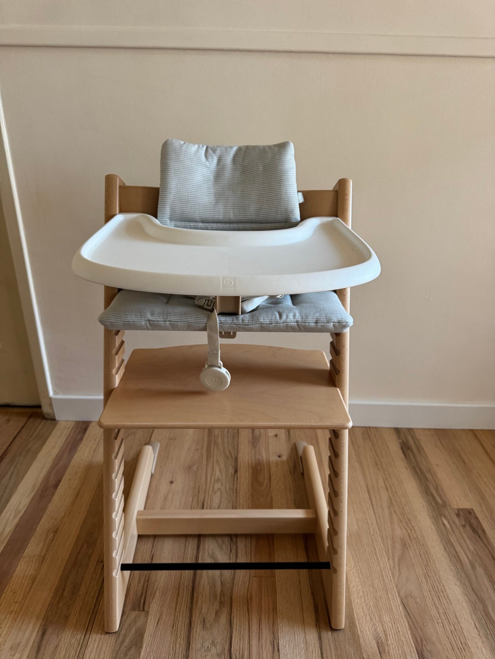 A wooden high chair with a gray cushion and a white plastic tray is placed on a wooden floor against a light-colored wall.