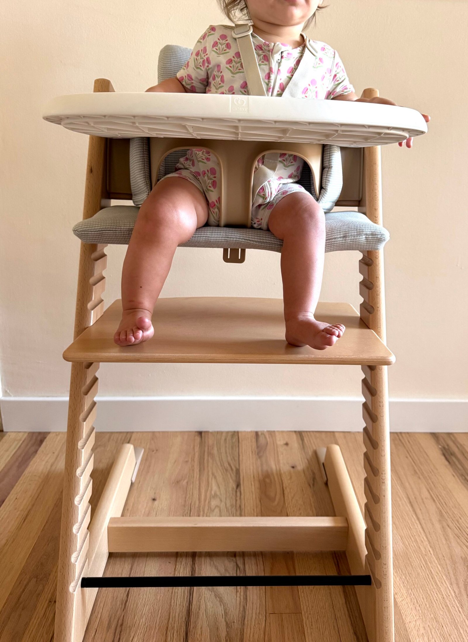 A baby wearing a floral outfit sits in a wooden high chair with feet dangling, tray attached, and a safety harness fastened.
