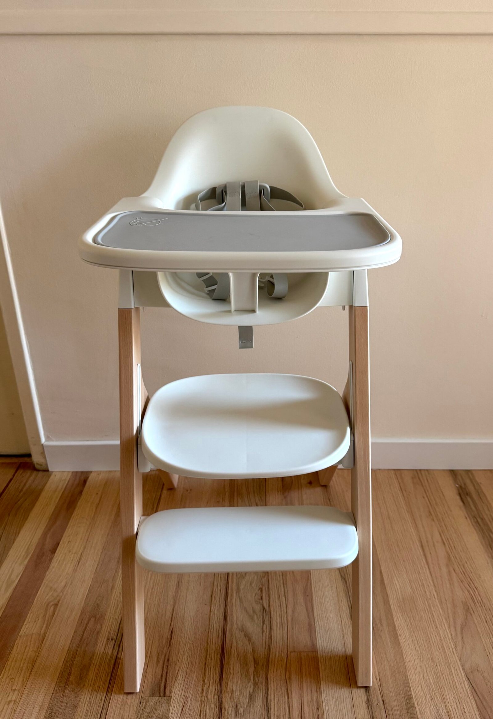 A white and light wood high chair with a gray tray is positioned on a wooden floor against a beige wall.