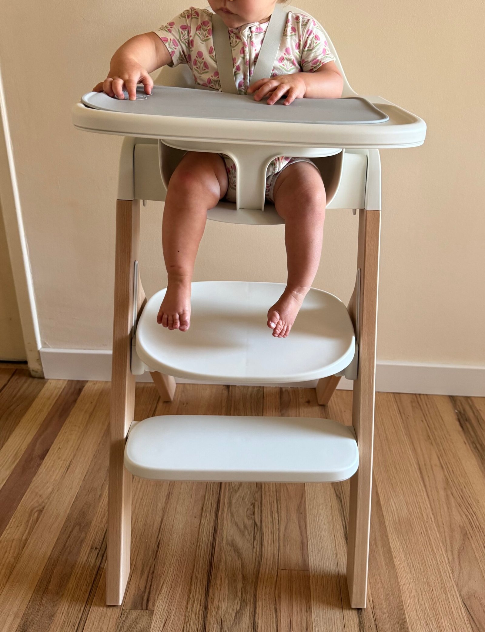 A baby sits in a modern high chair with a tray, wearing a patterned outfit. Only the baby’s torso, arms, and legs are visible. The chair is on a wooden floor against a plain wall.