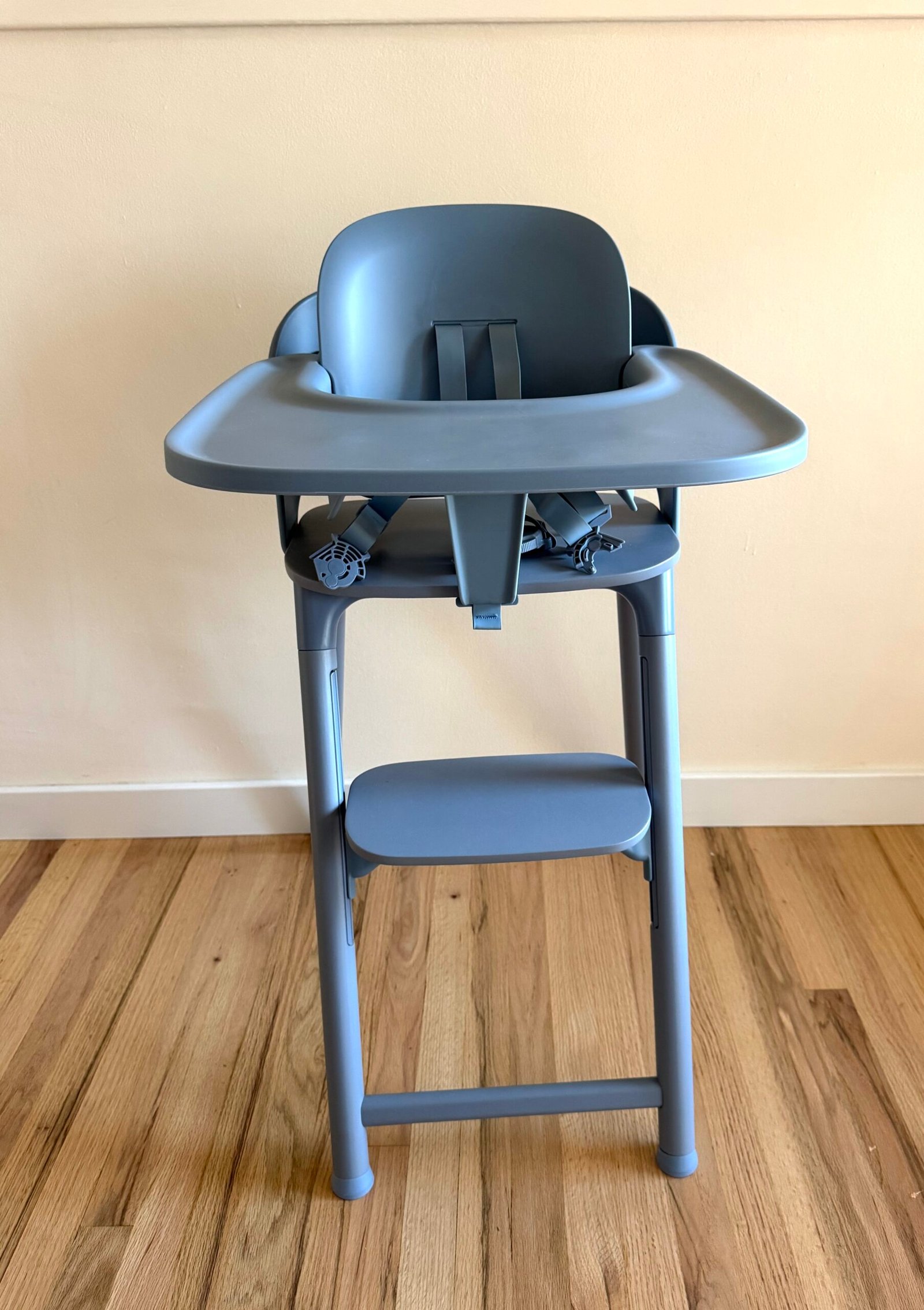 A modern gray high chair with tray and safety harness stands on a wooden floor against a plain light-colored wall.