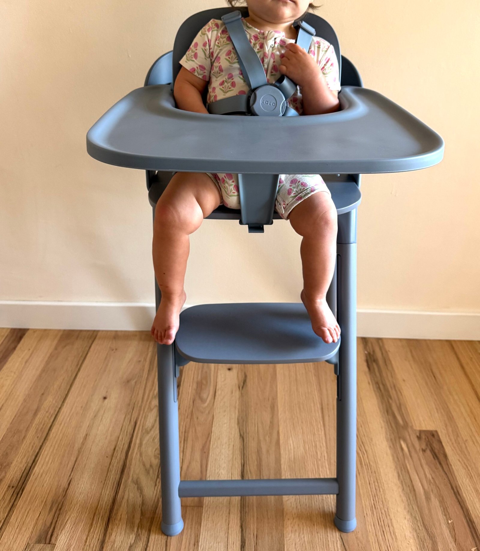 A baby wearing a floral onesie sits in a gray high chair with a safety harness, feet resting on the footrest, against a light wall and wooden floor.