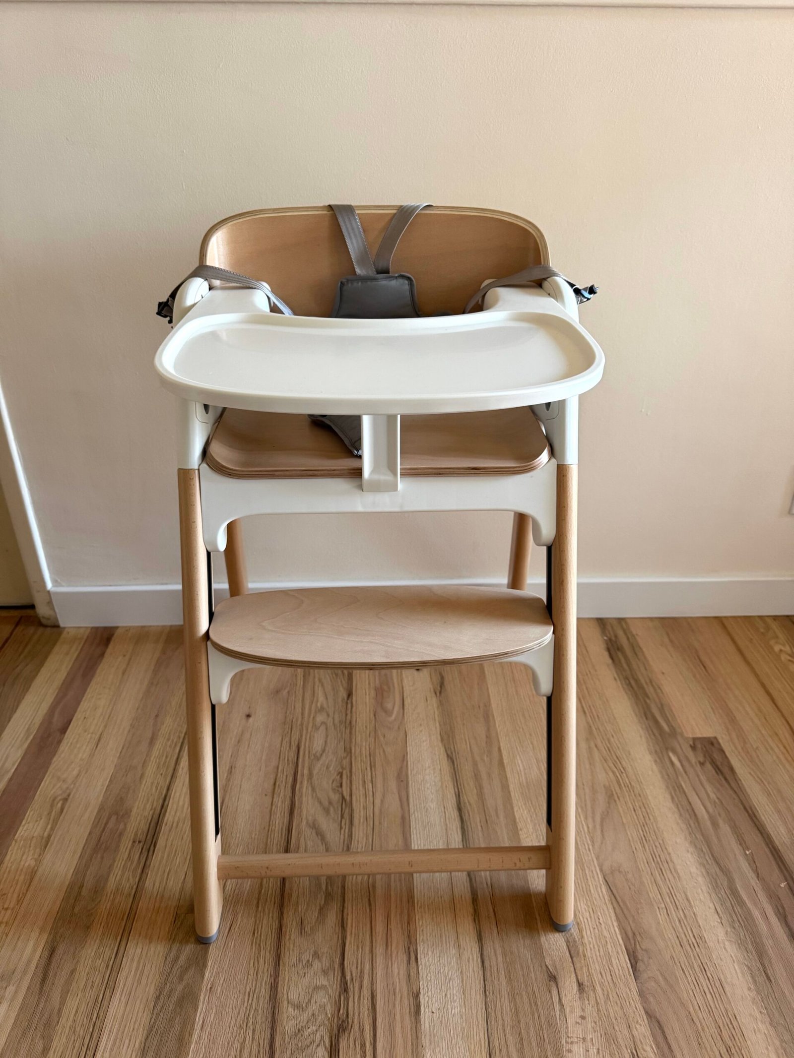 A wooden high chair with a white plastic tray and a gray safety harness is placed on a wooden floor against a light-colored wall.